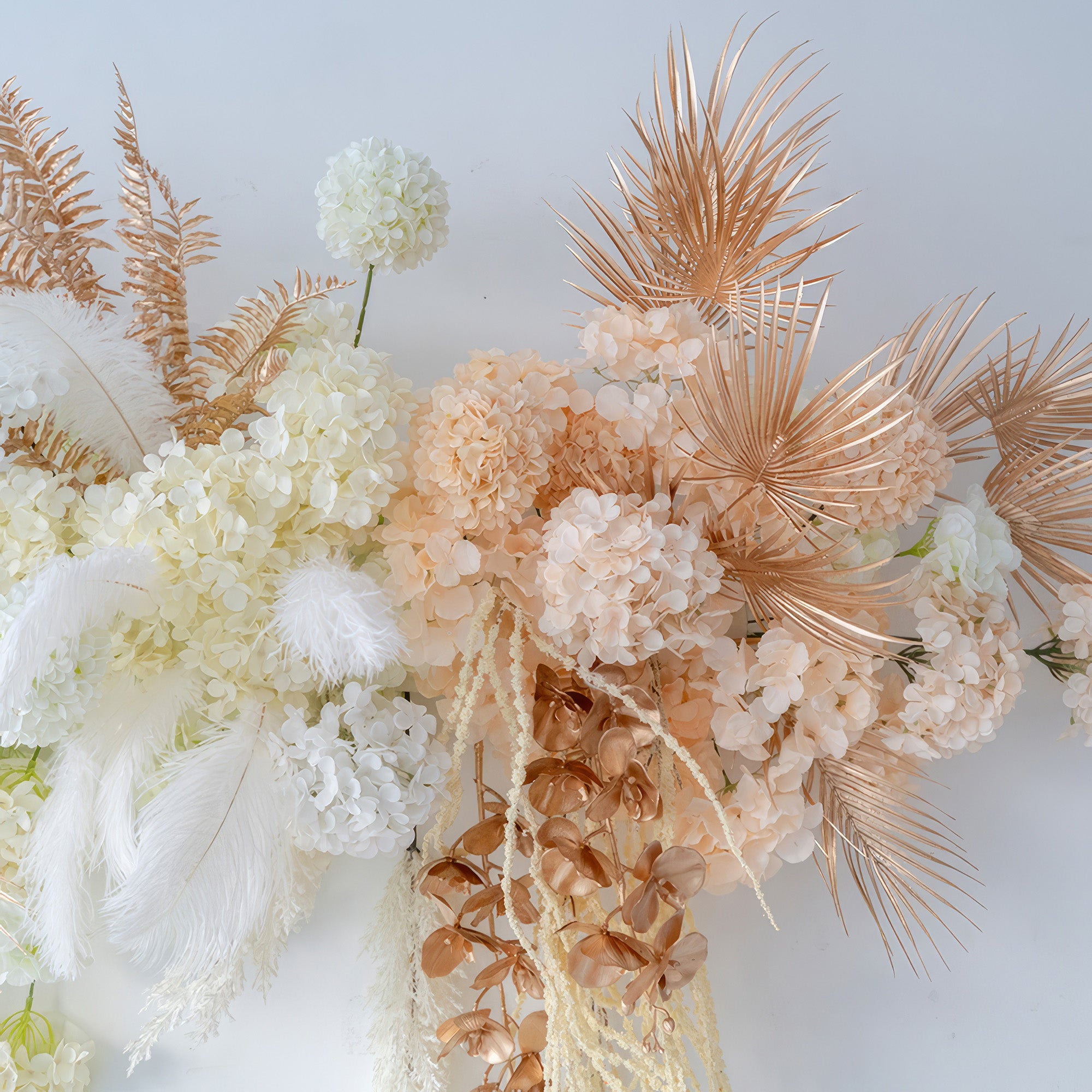 This image shows an artificial flower arrangement with hydrangeas as the main flower, along with palm - like fronds and feathers. Colors include white, peach, and metallic gold, creating a luxurious and modern style.