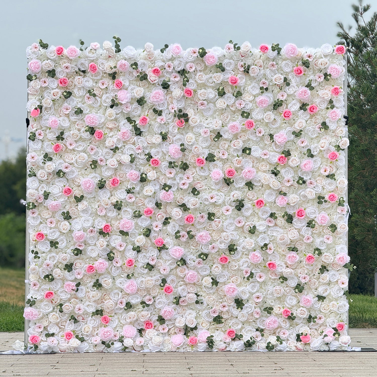 This photo showcases a stunning floral wall, likely by ValarFlowers, featuring artificial roses in soft pink and white hues, interspersed with greenery. The dense arrangement creates a romantic and elegant ambiance, perfect for weddings or other formal events. The realistic appearance of the faux flowers adds a touch of timeless beauty, making it an ideal backdrop for capturing memorable moments with a dreamy, celebratory atmosphere.