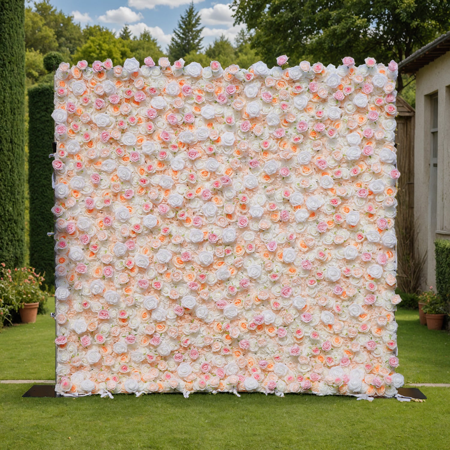 This photo showcases a large floral wall, likely by ValarFlowers, set in an outdoor garden. The wall is densely covered with artificial roses in soft hues of white, pink, and peach, creating a romantic, elegant aesthetic. The lush greenery and distant trees in the background enhance the natural, serene ambiance. This setup is perfect for weddings, photoshoots, or events, adding a touch of timeless beauty and sophistication to any occasion. The realistic design of the faux flowers ensures long-lasting charm.