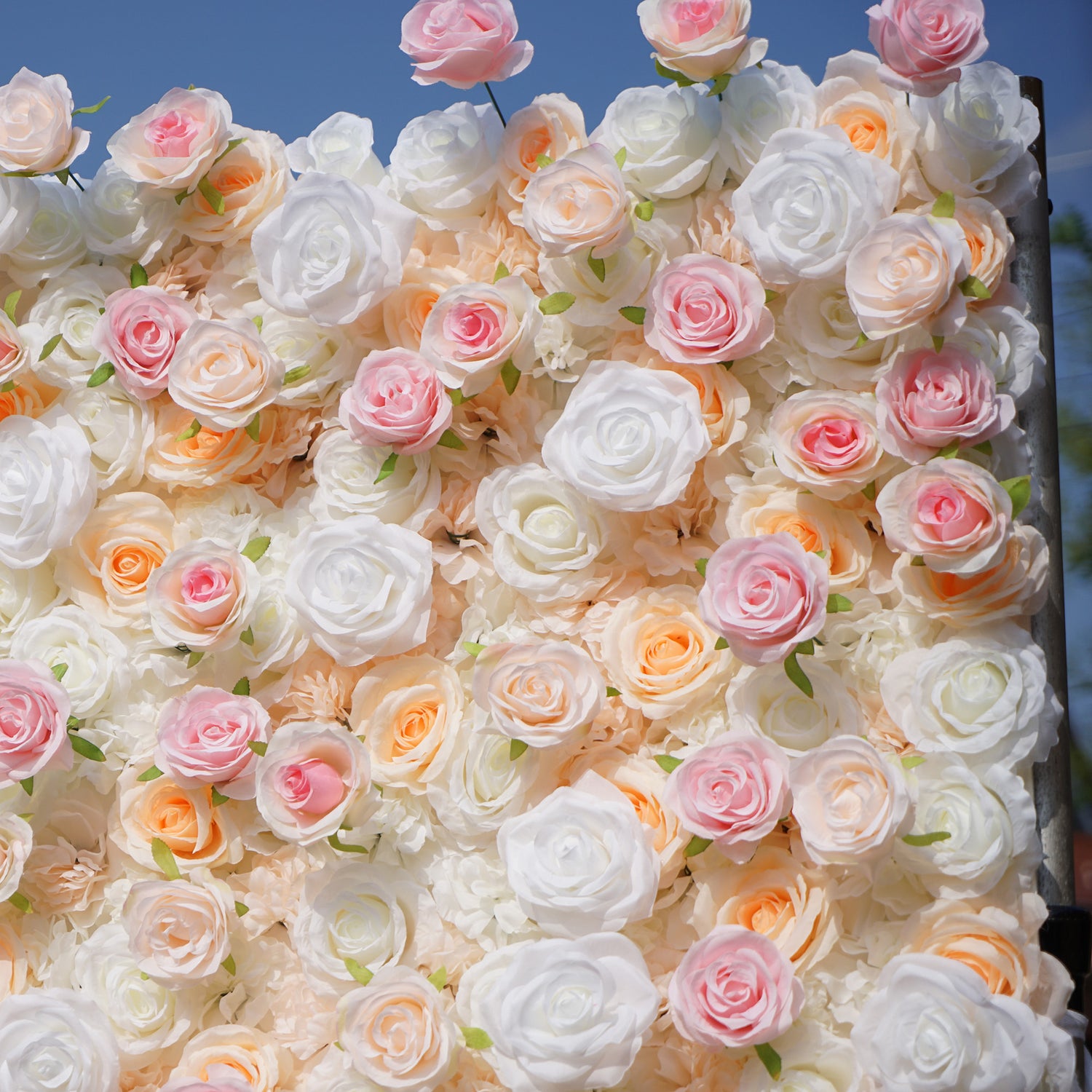 This photo captures a stunning floral wall, likely by ValarFlowers, brimming with artificial roses in soft shades of white, pink, and peach. The dense arrangement creates a romantic, dreamy ambiance, perfect for weddings or events. The realistic petals and lush green accents enhance the natural look, while the bright blue sky in the background adds a touch of serenity. This setup offers long-lasting beauty, ideal for creating memorable moments and elegant backdrops.