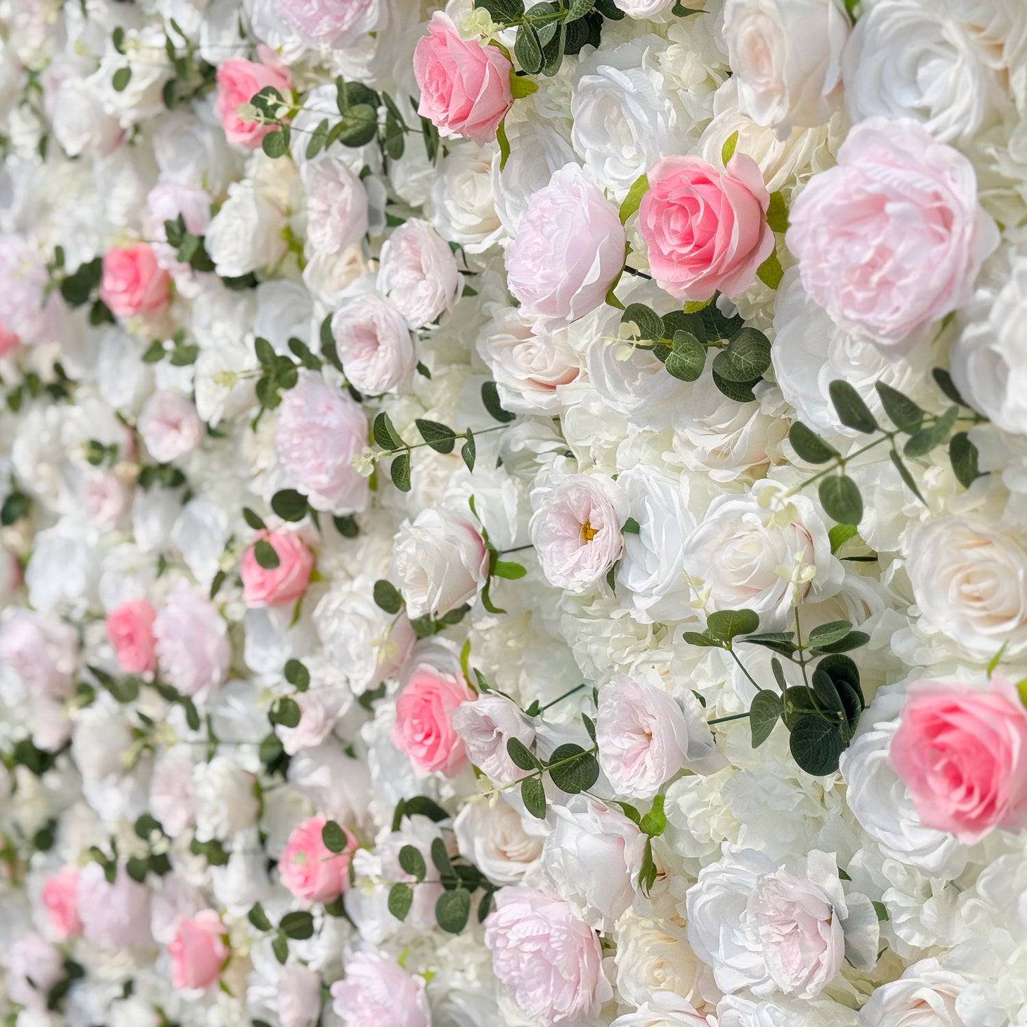 This photo showcases a stunning floral wall, likely by ValarFlowers, featuring artificial roses in soft pink and white hues, interspersed with greenery. The dense arrangement creates a romantic and elegant ambiance, perfect for weddings or other formal events. The realistic appearance of the faux flowers adds a touch of timeless beauty, making it an ideal backdrop for capturing memorable moments with a dreamy, celebratory atmosphere.