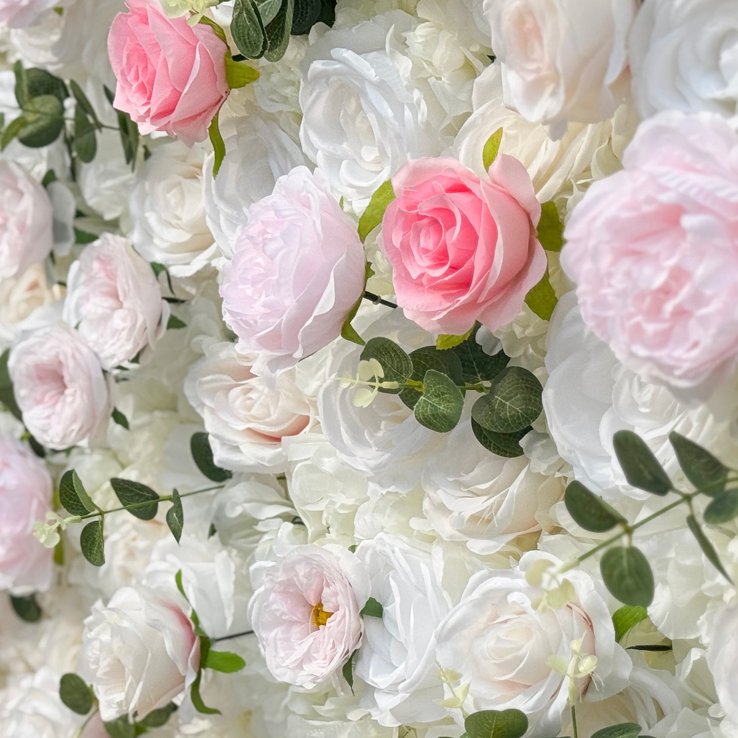 This photo showcases a stunning floral wall, likely by ValarFlowers, featuring artificial roses in soft pink and white hues, interspersed with greenery. The dense arrangement creates a romantic and elegant ambiance, perfect for weddings or other formal events. The realistic appearance of the faux flowers adds a touch of timeless beauty, making it an ideal backdrop for capturing memorable moments with a dreamy, celebratory atmosphere.