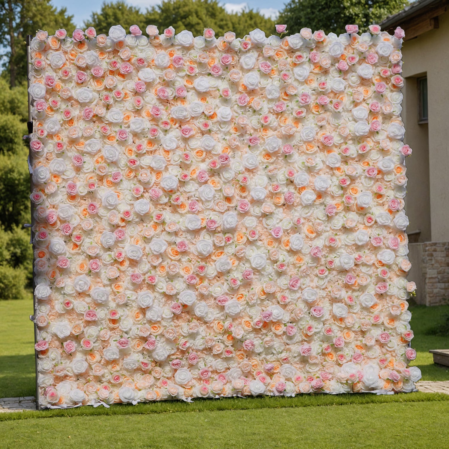 This photo showcases a large floral wall, likely by ValarFlowers, set in an outdoor garden. The wall is densely covered with artificial roses in soft hues of white, pink, and peach, creating a romantic, elegant aesthetic. The lush greenery and distant trees in the background enhance the natural, serene ambiance. This setup is perfect for weddings, photoshoots, or events, adding a touch of timeless beauty and sophistication to any occasion. The realistic design of the faux flowers ensures long-lasting charm.