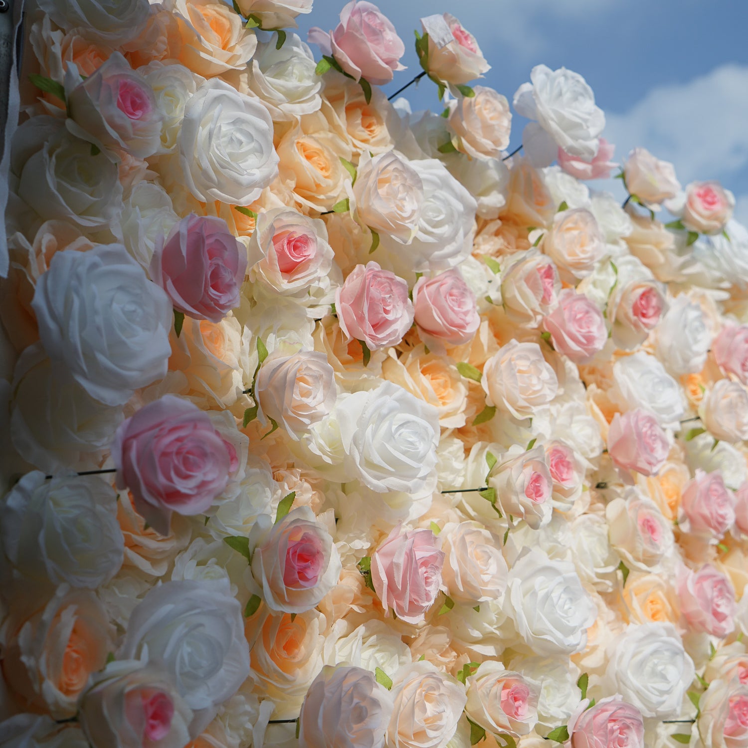 This photo showcases a lush floral wall composed of artificial roses, likely by ValarFlowers. The blooms come in soft shades of white, peach, and pink, creating a romantic and elegant aesthetic. This setup is perfect for events like weddings, where the delicate hues and realistic appearance of the faux flowers add a touch of timeless beauty. The dense arrangement and pastel colors evoke a dreamy, celebratory atmosphere, making it an ideal backdrop for capturing memorable moments.