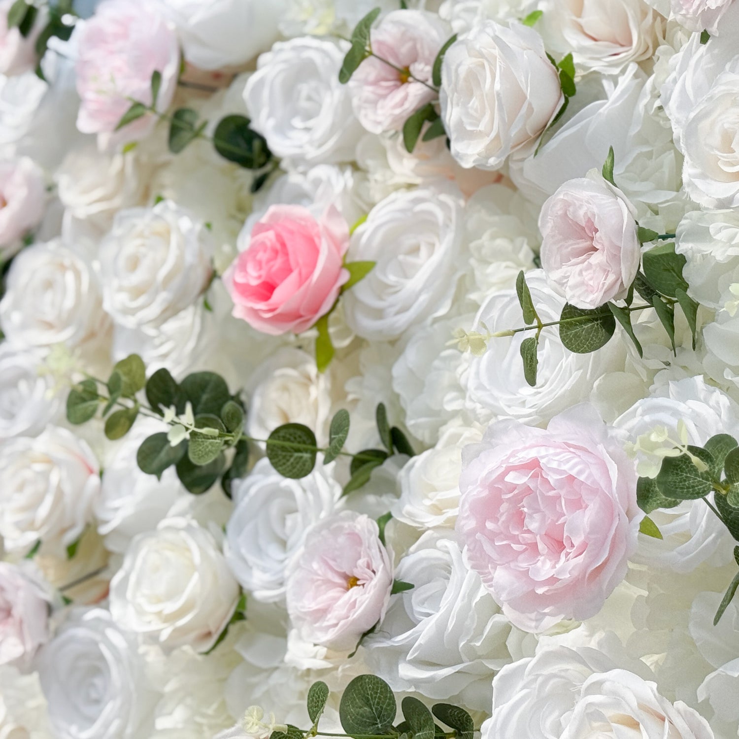 This photo showcases a stunning floral wall, likely by ValarFlowers, featuring artificial roses in soft pink and white hues, interspersed with greenery. The dense arrangement creates a romantic and elegant ambiance, perfect for weddings or other formal events. The realistic appearance of the faux flowers adds a touch of timeless beauty, making it an ideal backdrop for capturing memorable moments with a dreamy, celebratory atmosphere.