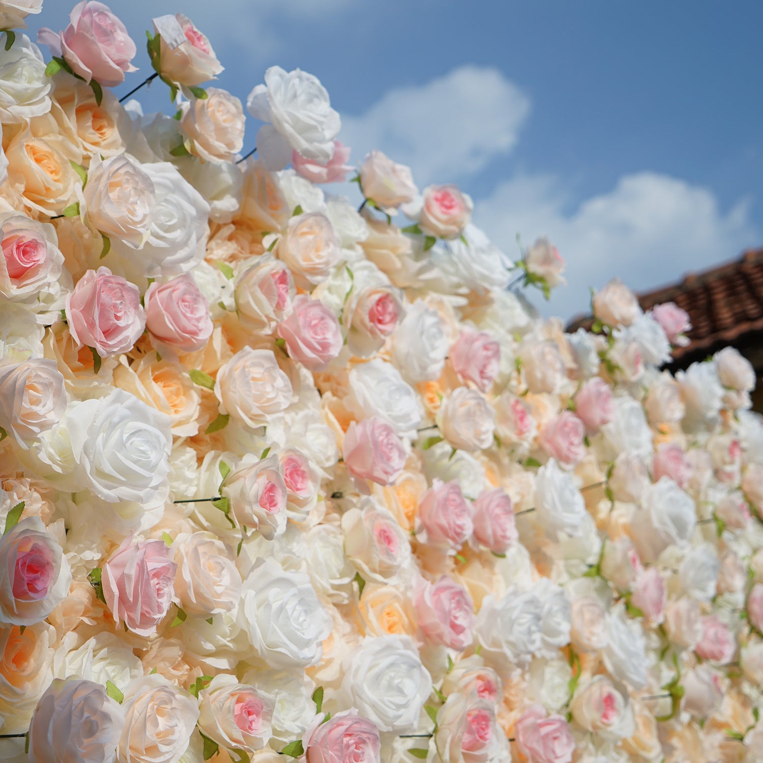 This photo showcases a lush floral wall composed of artificial roses, likely by ValarFlowers. The blooms come in soft shades of white, peach, and pink, creating a romantic and elegant aesthetic. This setup is perfect for events like weddings, where the delicate hues and realistic appearance of the faux flowers add a touch of timeless beauty. The dense arrangement and pastel colors evoke a dreamy, celebratory atmosphere, making it an ideal backdrop for capturing memorable moments.