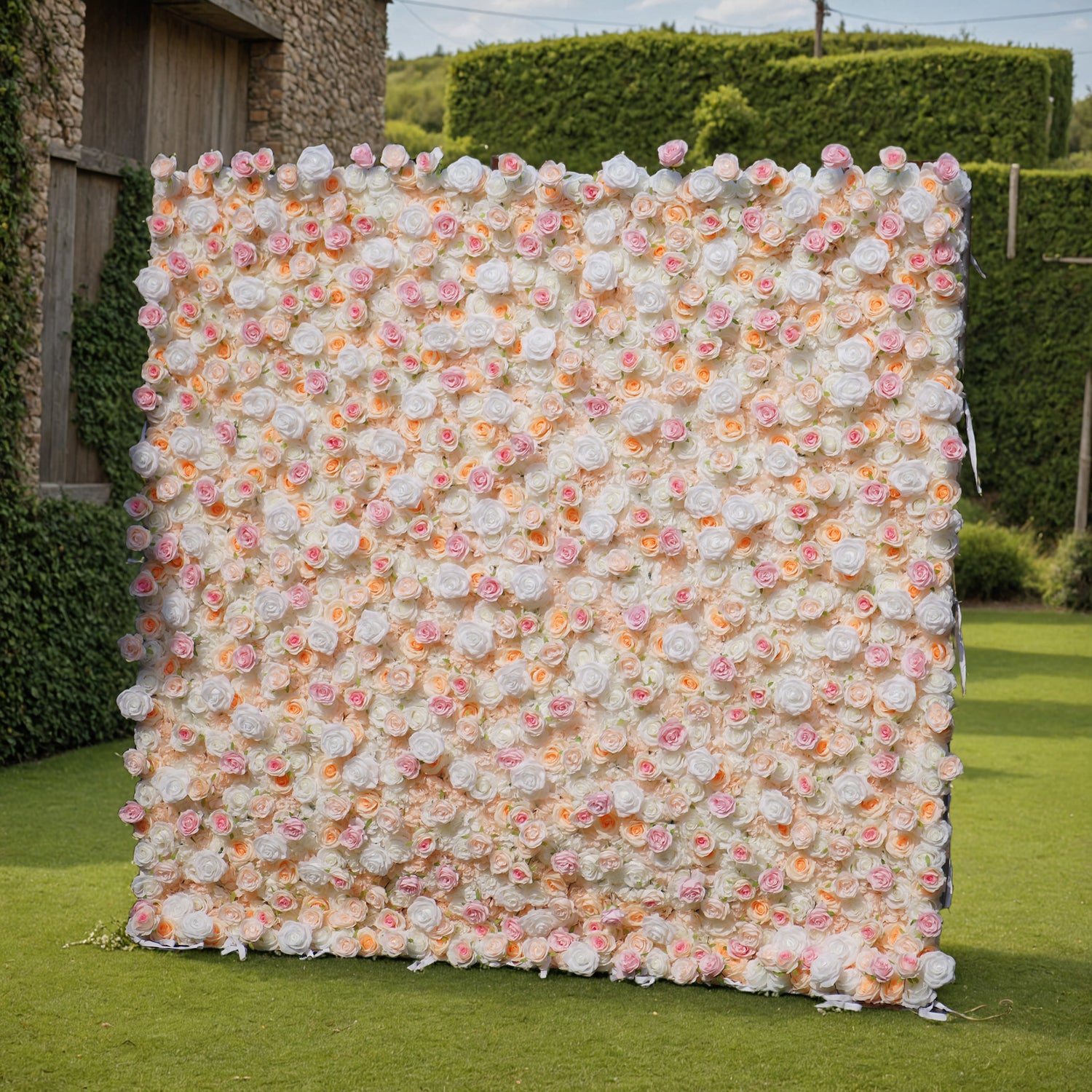 This photo showcases a large floral wall, likely by ValarFlowers, set in an outdoor garden. The wall is densely covered with artificial roses in soft hues of white, pink, and peach, creating a romantic, elegant aesthetic. The lush greenery and distant trees in the background enhance the natural, serene ambiance. This setup is perfect for weddings, photoshoots, or events, adding a touch of timeless beauty and sophistication to any occasion. The realistic design of the faux flowers ensures long-lasting charm.