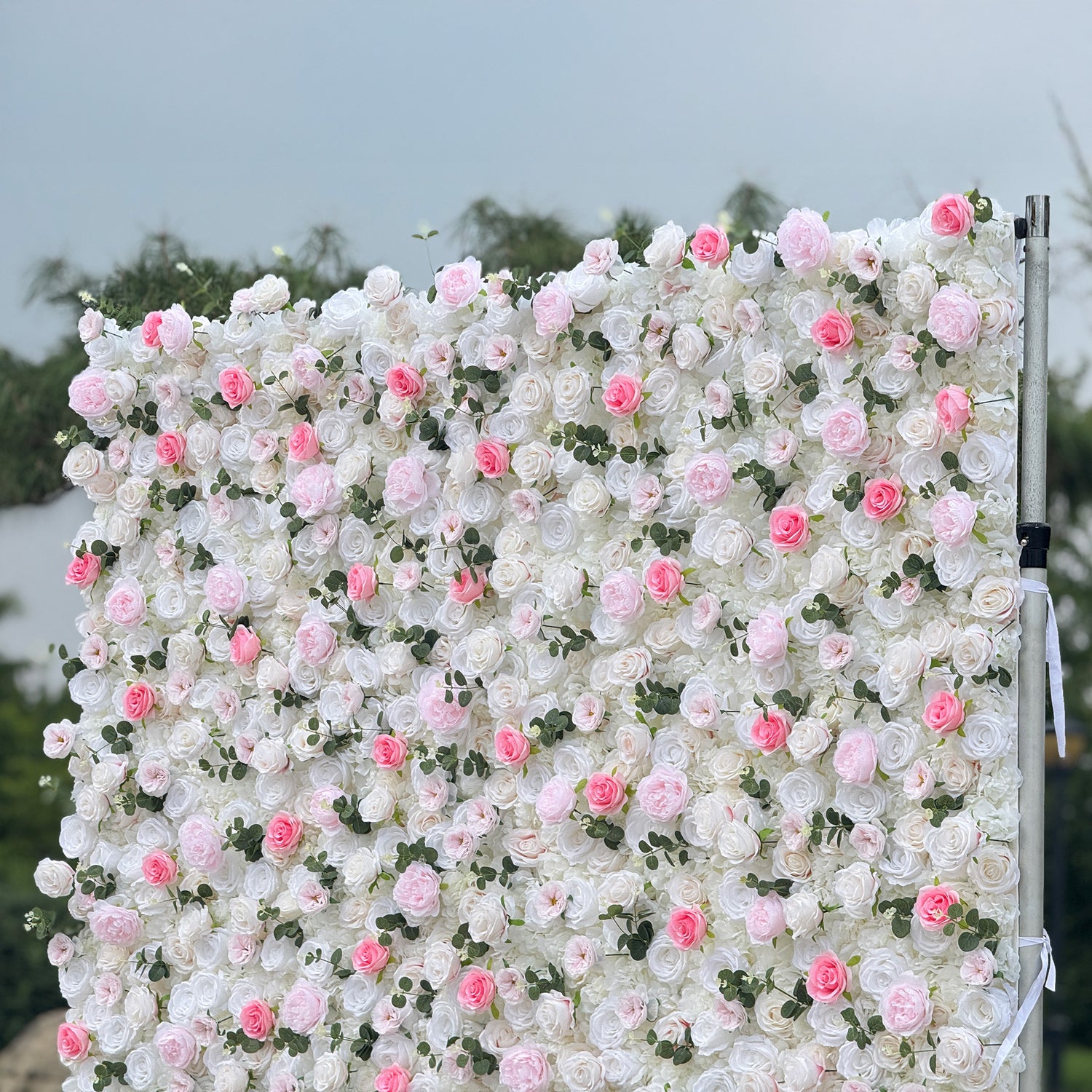 This photo showcases a stunning floral wall, likely by ValarFlowers, featuring artificial roses in soft pink and white hues, interspersed with greenery. The dense arrangement creates a romantic and elegant ambiance, perfect for weddings or other formal events. The realistic appearance of the faux flowers adds a touch of timeless beauty, making it an ideal backdrop for capturing memorable moments with a dreamy, celebratory atmosphere.