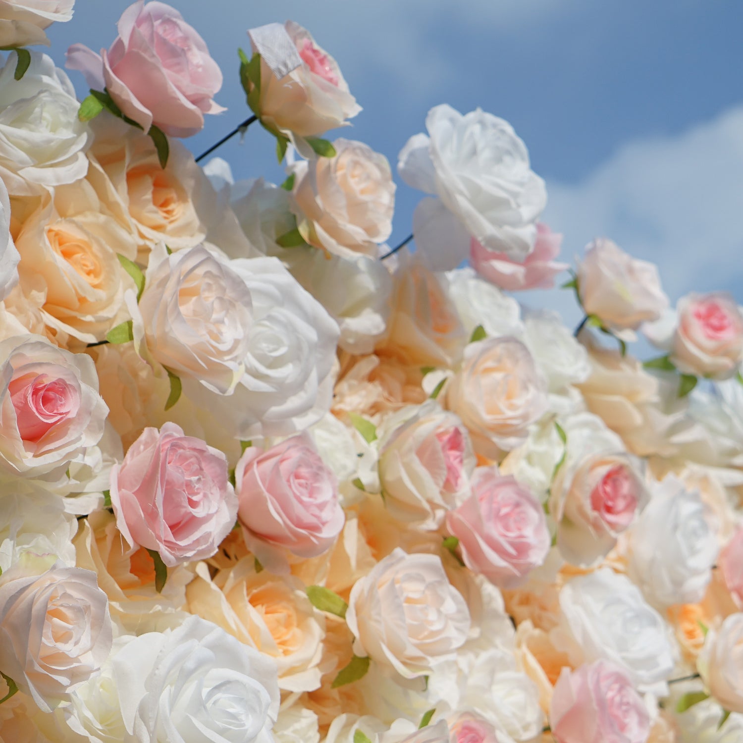 This photo showcases a lush floral wall composed of artificial roses, likely by ValarFlowers. The blooms come in soft shades of white, peach, and pink, creating a romantic and elegant aesthetic. This setup is perfect for events like weddings, where the delicate hues and realistic appearance of the faux flowers add a touch of timeless beauty. The dense arrangement and pastel colors evoke a dreamy, celebratory atmosphere, making it an ideal backdrop for capturing memorable moments.