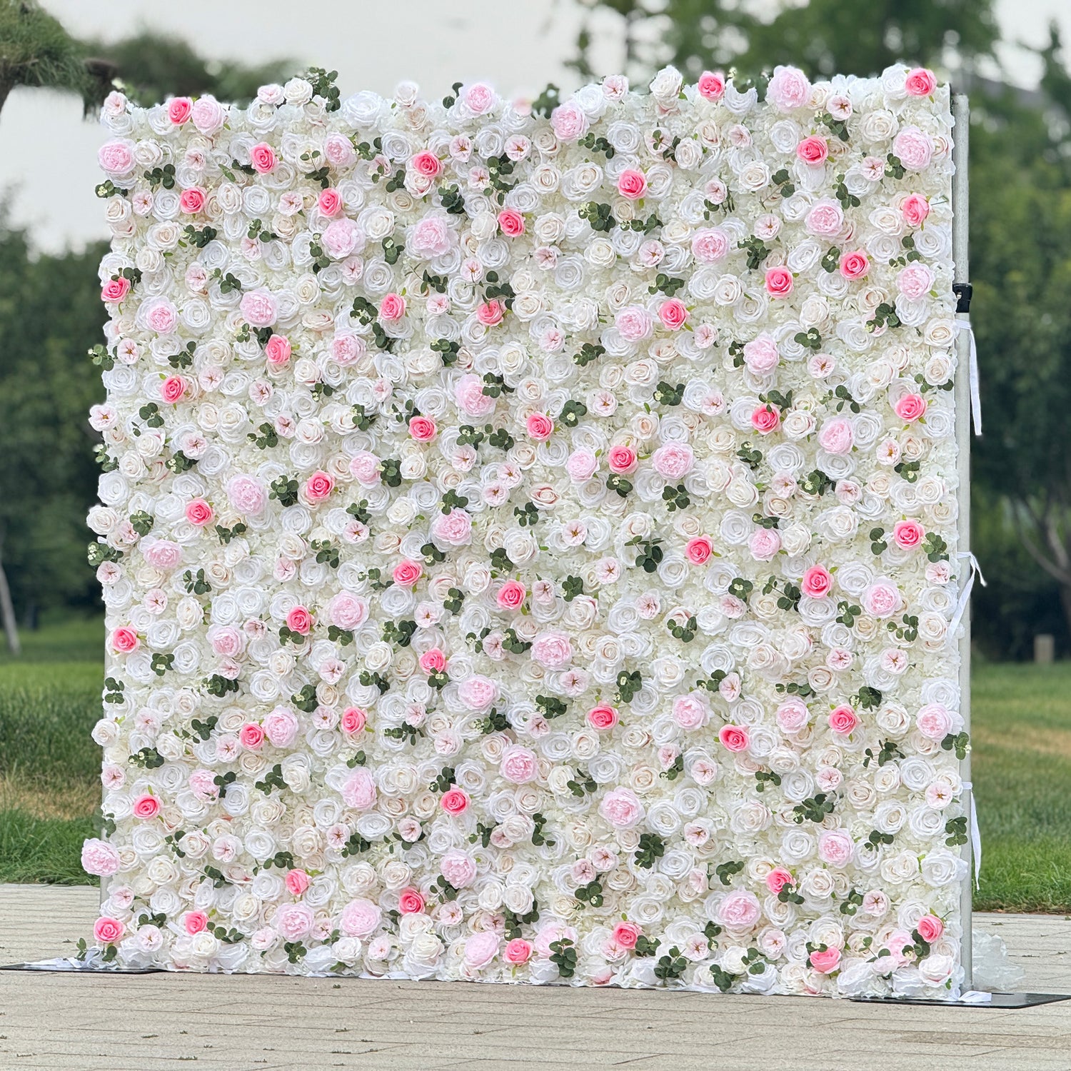 This photo showcases a stunning floral wall, likely by ValarFlowers, featuring artificial roses in soft pink and white hues, interspersed with greenery. The dense arrangement creates a romantic and elegant ambiance, perfect for weddings or other formal events. The realistic appearance of the faux flowers adds a touch of timeless beauty, making it an ideal backdrop for capturing memorable moments with a dreamy, celebratory atmosphere.