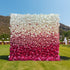 This image showcases a striking flower wall made of artificial flowers, set outdoors on a grassy area. The wall features a gradient design transitioning from white at the top to deep pink at the bottom, primarily composed of rose - like blooms. The color scheme creates a visually appealing ombre effect, exuding a romantic and elegant style, perfect for events such as weddings or photo - shoots.