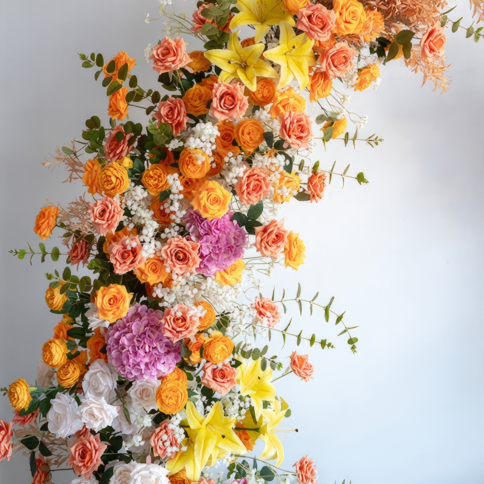 This is a strikingly colorful floral arrangement, likely part of an arch. It combines a variety of flowers including orange and peach roses, vivid yellow lilies, large purple hydrangeas, and white blooms. Green foliage and some dried elements add texture. The rich palette creates a lively, celebratory look, perfect for weddings or festive events.
