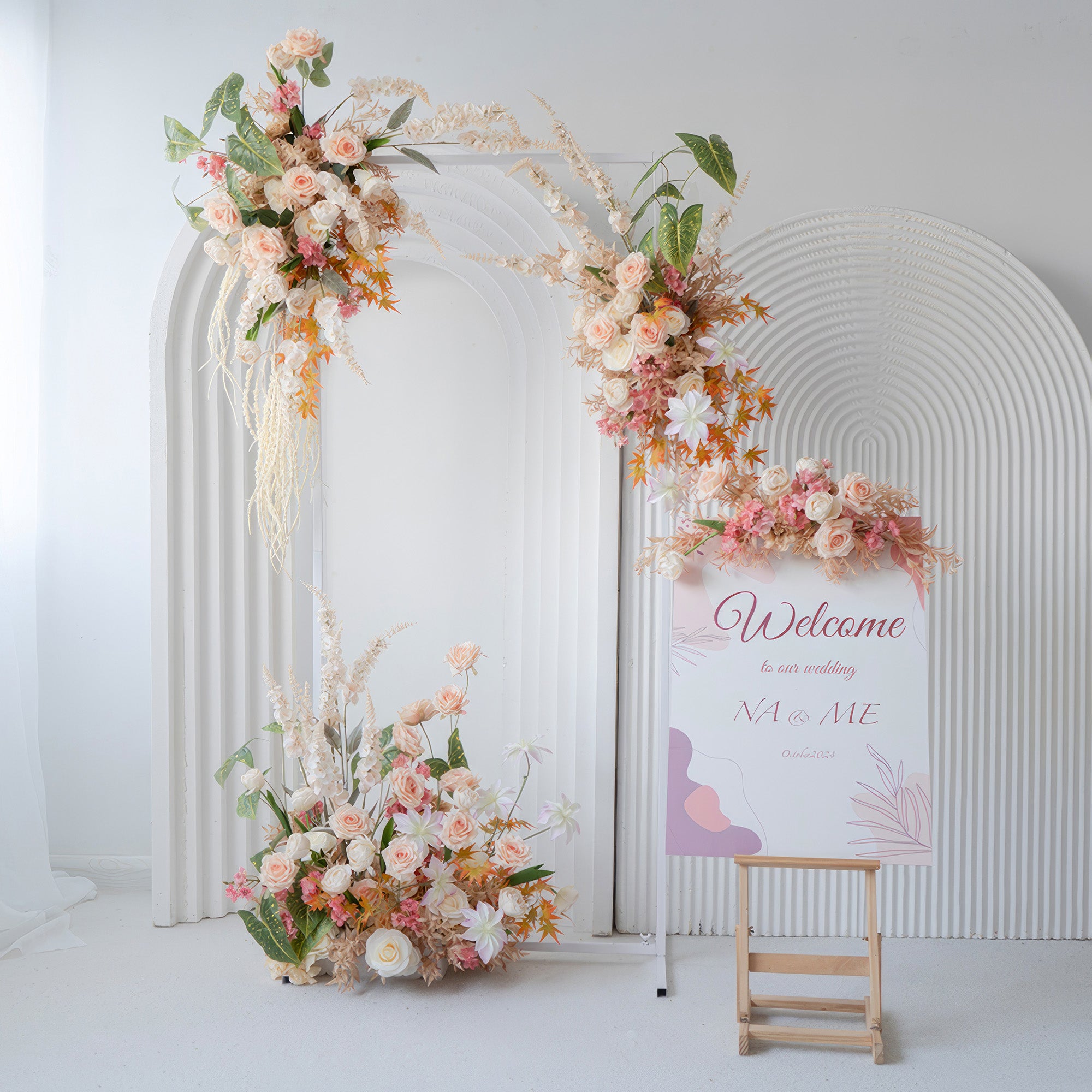 The image shows a wedding - themed decor with an arch and ground floral arrangements. Featuring roses and other blooms in soft peaches, pinks, and whites, along with a “Welcome” sign, it has a warm, romantic style.