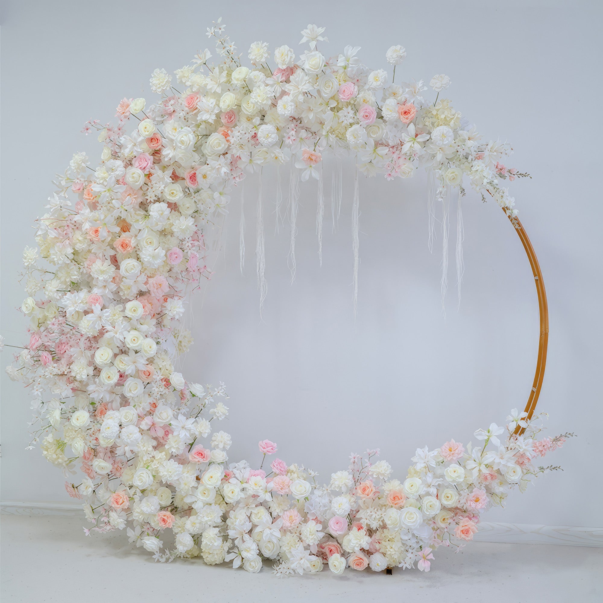 This image shows a circular floral arch adorned with artificial flowers. Colors are soft white, light pink, and peach. Flowers include roses and smaller blooms. The style is elegant and romantic, suitable for a wedding.