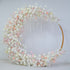 This image shows a circular floral arch adorned with artificial flowers. Colors are soft white, light pink, and peach. Flowers include roses and smaller blooms. The style is elegant and romantic, suitable for a wedding.