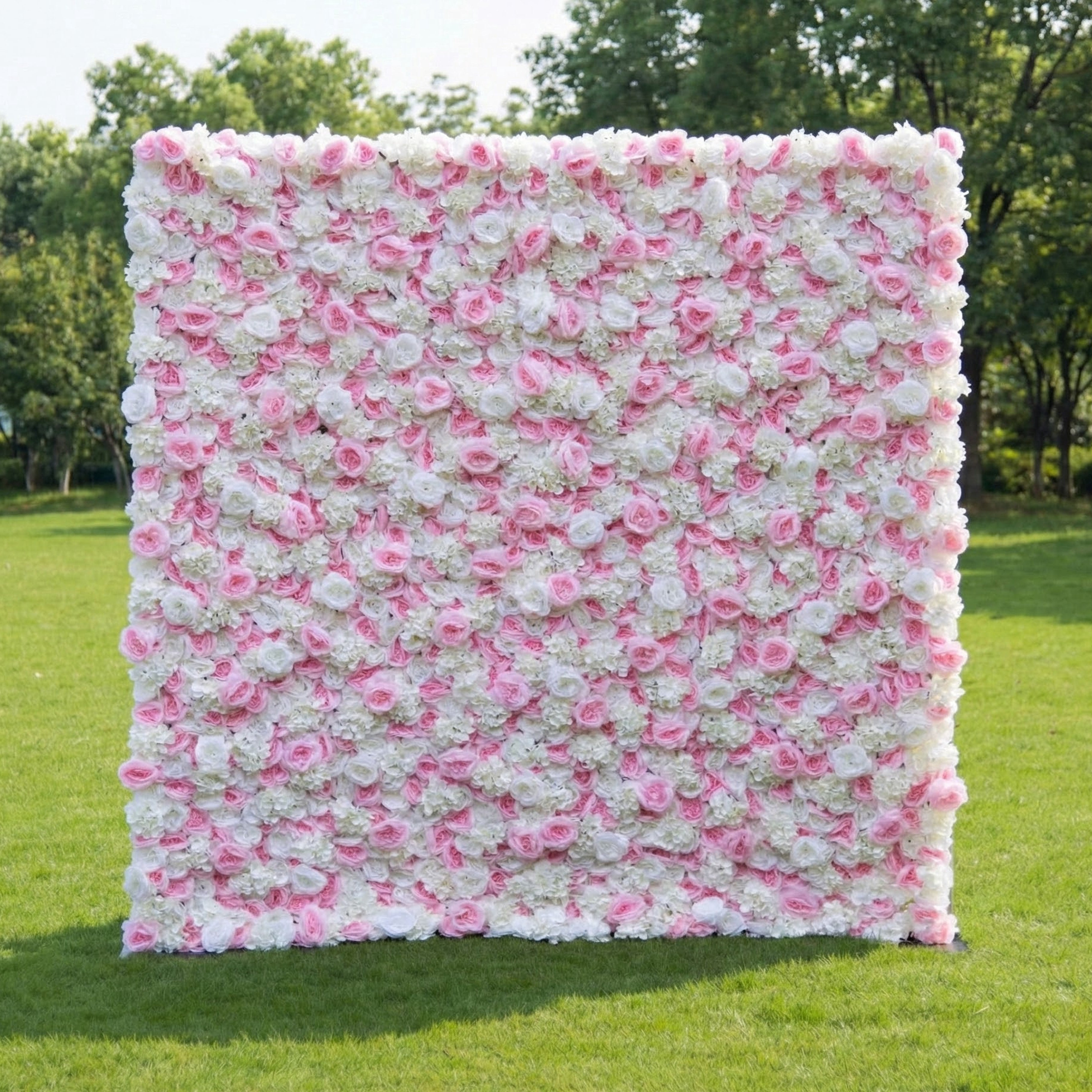 This outdoor floral backdrop (set on lush green grass, tree-framed) is a dense wall of soft pink roses and white filler blooms, creating an airy, romantic texture. The delicate, blush-and-ivory palette exudes gentle elegance, making it ideal for garden weddings, baby showers, or intimate outdoor events that lean into timeless, feminine charm.