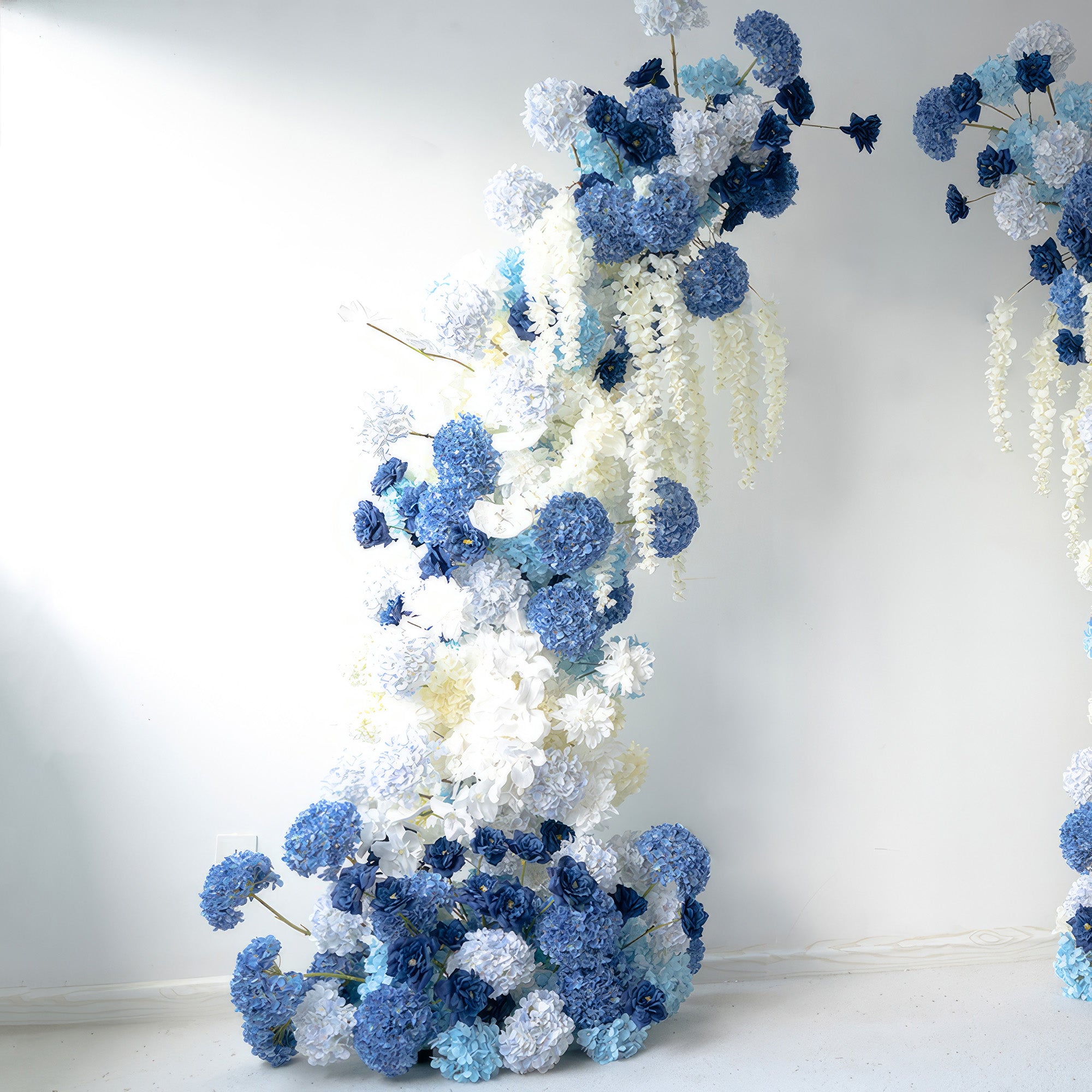 This floral arrangement features a beautiful blend of deep and light blues with pure whites. Dominated by hydrangeas and accented with hanging flowers, it offers a serene and elegant style. Ideal for weddings, events, or as a photo backdrop, it brings a touch of tranquility and beauty.