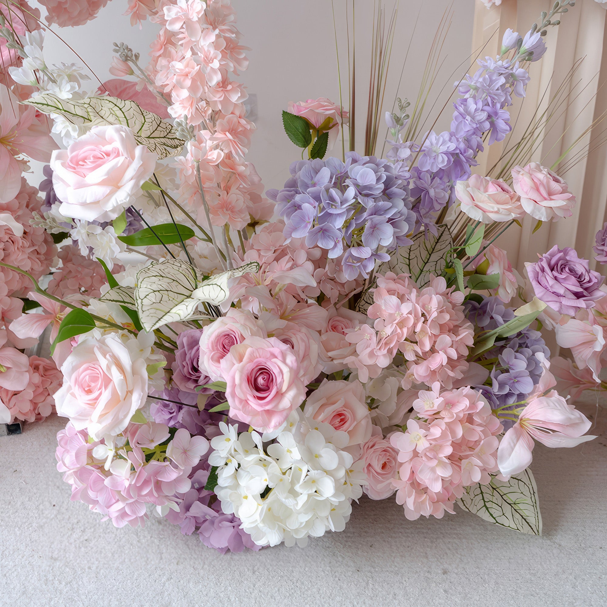 This is a beautiful floral arrangement with a soft pastel color palette. It includes pink roses, lavender and white hydrangeas, along with some leaves that have distinct veining. The combination of these artificial flowers creates a romantic and elegant look, suitable for wedding decorations, bridal showers, or other delicate and charming events.