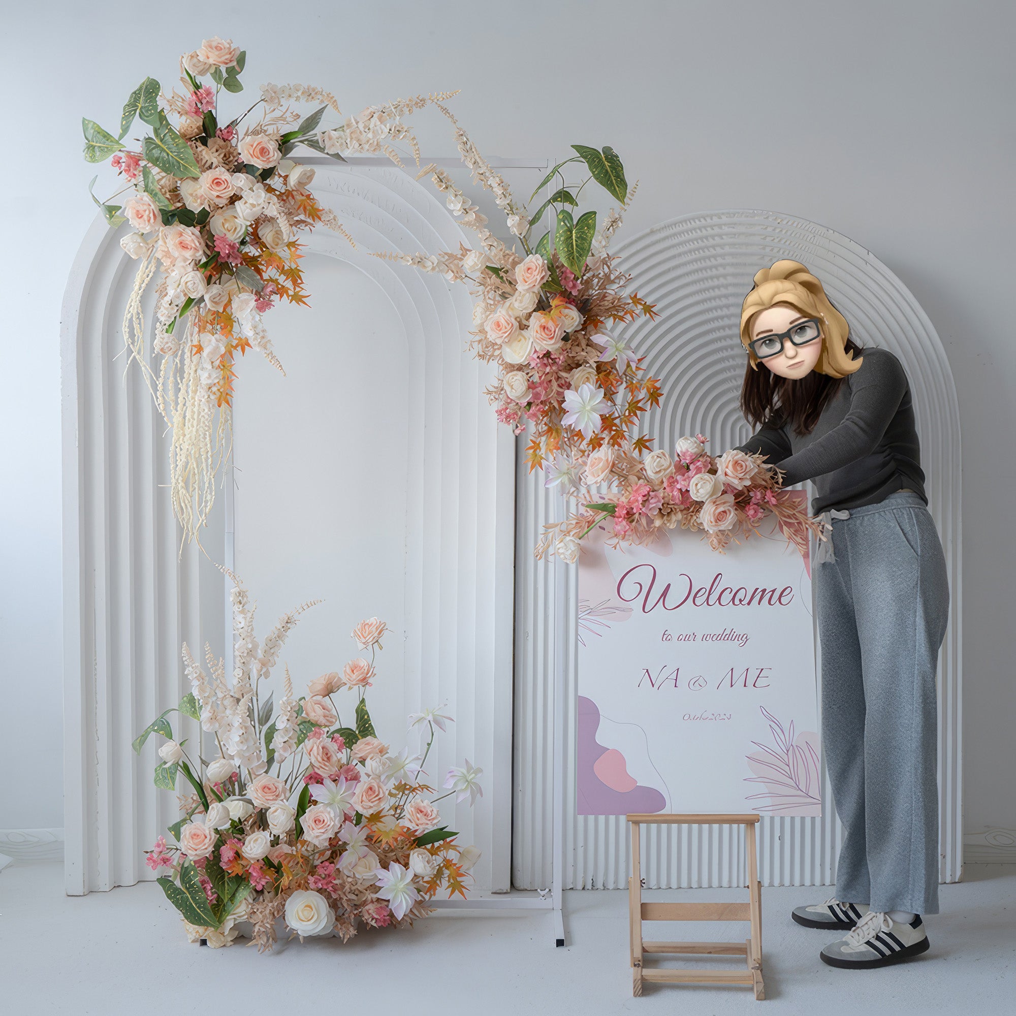The image depicts a wedding - themed decor with an arch covered in artificial roses, daisies, and greenery. Colors are soft pink, peach, and white. A "Welcome" sign is present, and the overall style is warm and inviting.
