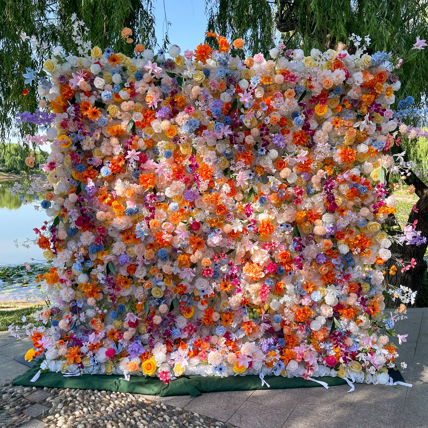 This image features a lush floral wall outdoors by a body of water, surrounded by greenery. The wall is densely packed with a variety of artificial flowers in a vibrant color palette, including orange, pink, purple, blue, yellow, and white. Flower types appear to include roses, daisies, and hydrangea - like blooms. The overall style is exuberant and cheerful, creating a lively and eye - catching display perfect for photo opportunities.