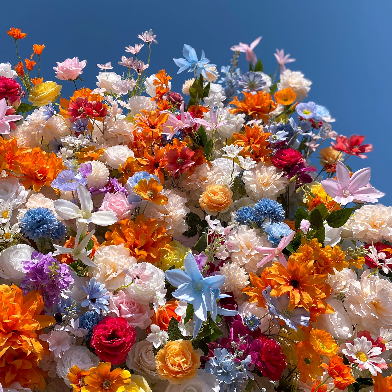 This image bursts with a vibrant array of artificial flowers set against a clear blue sky. The flowers showcase a rich palette of colors including bright orange, soft pink, vivid red, delicate blue, and pure white. Varieties such as roses, daisies, and star - shaped blooms are intermingled. The overall style is lively and cheerful, creating a whimsical and eye - catching display.
