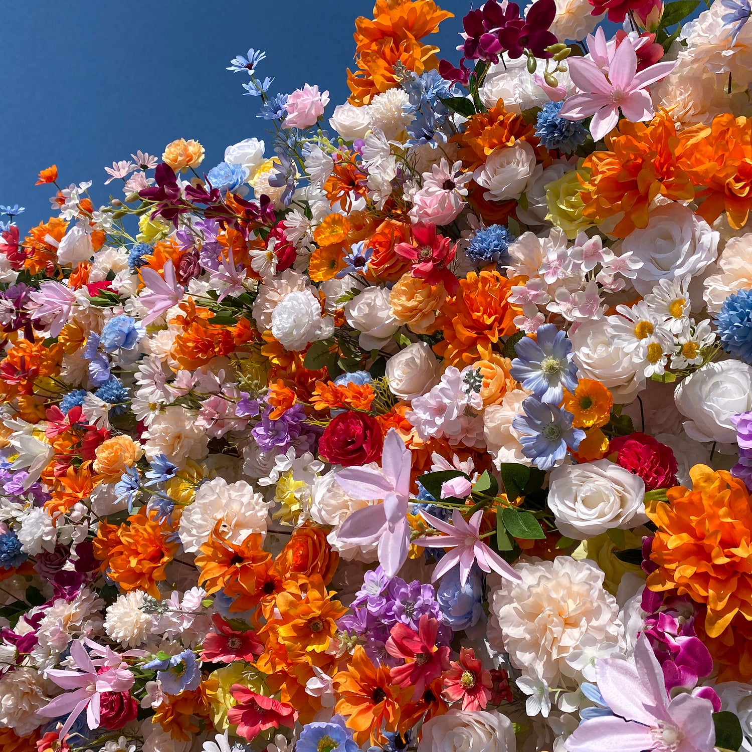 This image features a vibrant and densely packed arrangement of artificial flowers set against a clear blue sky. The flowers display a rich palette of colors, including bright oranges, soft pinks, deep purples, delicate blues, and crisp whites. Varieties such as roses, daisies, and star - shaped blooms are intermingled, creating a lively, cheerful, and whimsical style that exudes a sense of celebration.
