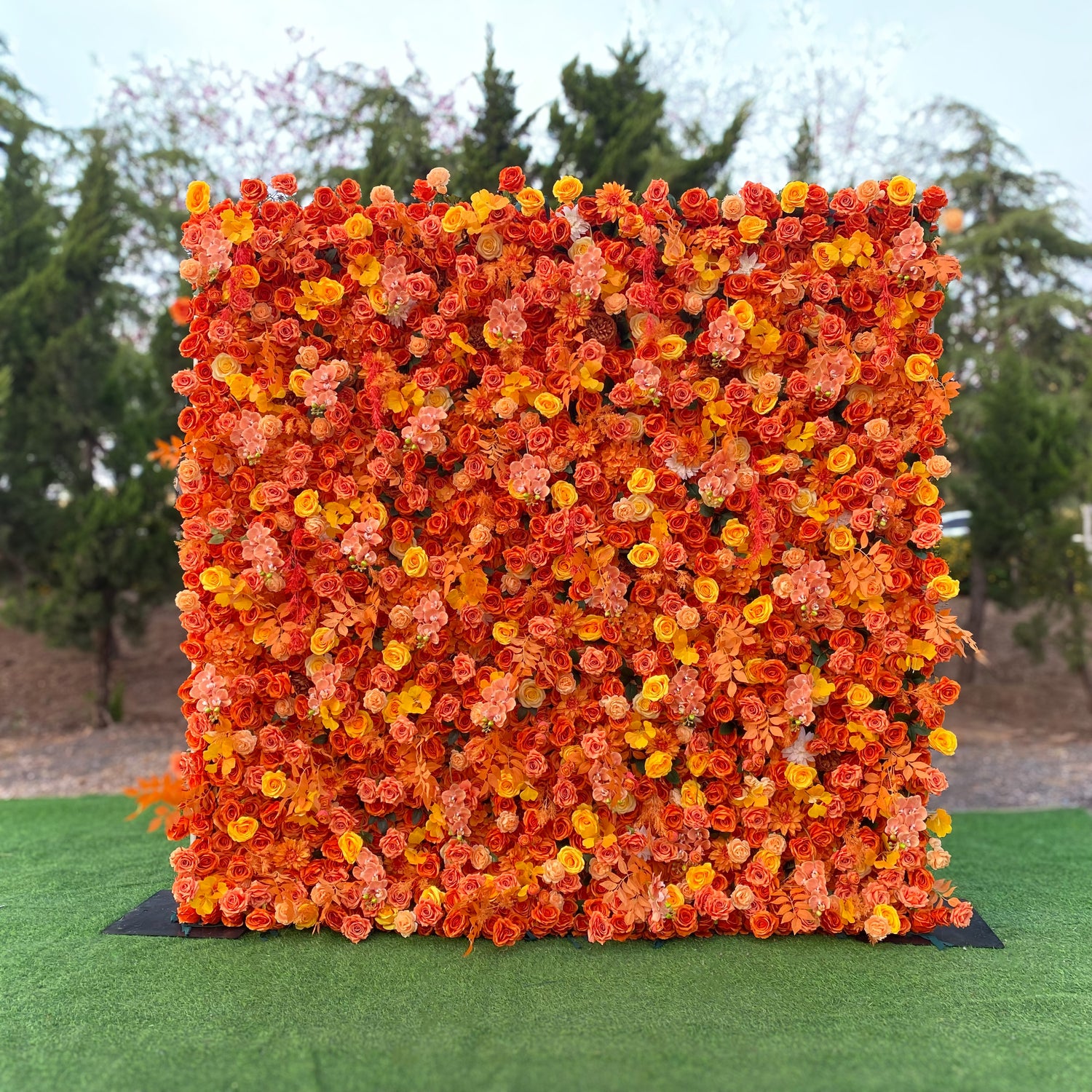 This image showcases a lush floral wall outdoors, set against a backdrop of greenery. The wall is densely adorned with artificial flowers in various shades of orange, from bright tangerine to soft peach, along with touches of yellow. Predominant flower types appear to be roses and small, clustered blooms. The overall style is vibrant and warm, evoking a sense of autumnal charm and creating a striking visual focal point.