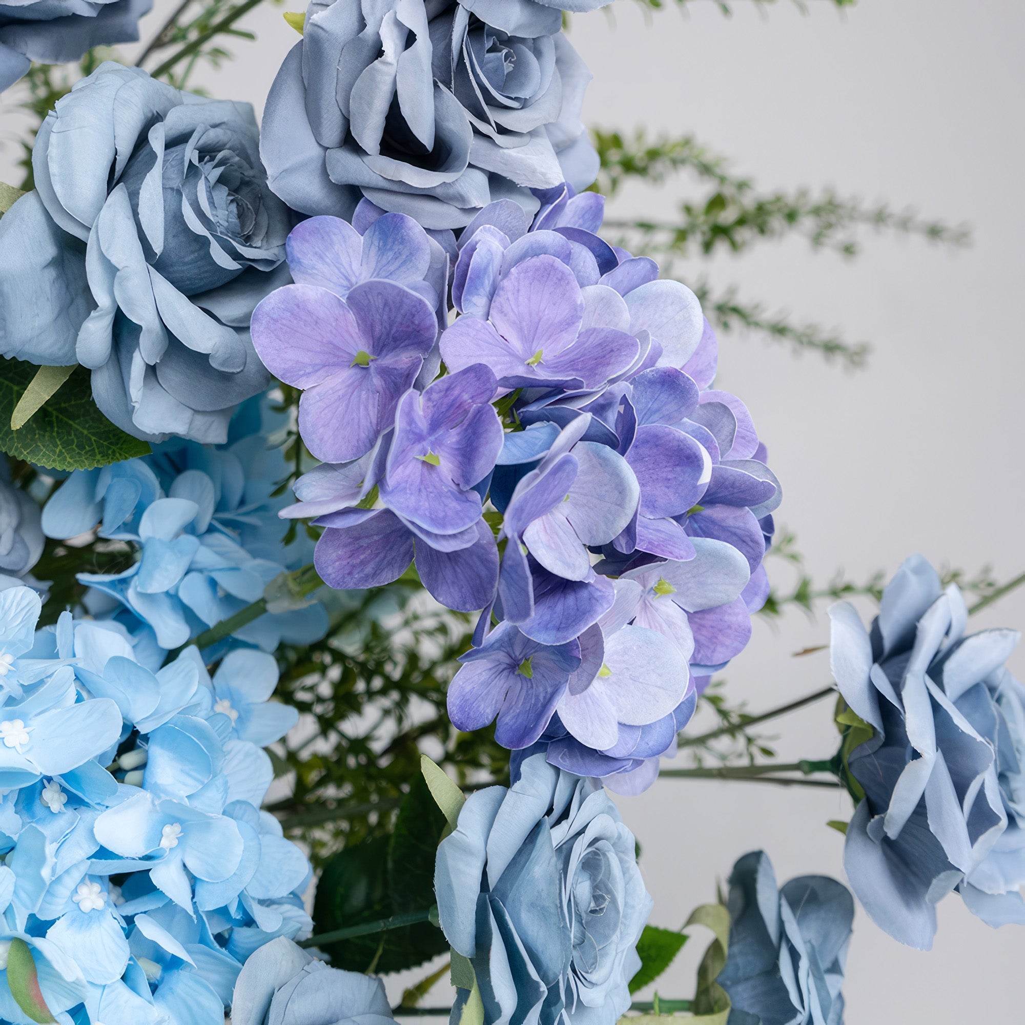 A close-up of a lush floral arrangement from Valar Flowers, featuring a harmonious blend of dusty blue roses, vibrant purple hydrangeas, and soft sky-blue blooms, intertwined with fresh green foliage. The rich, cool-toned palette and textured petals create a sophisticated, elegant aesthetic, ideal for wedding decor, centerpieces, or special event accents.