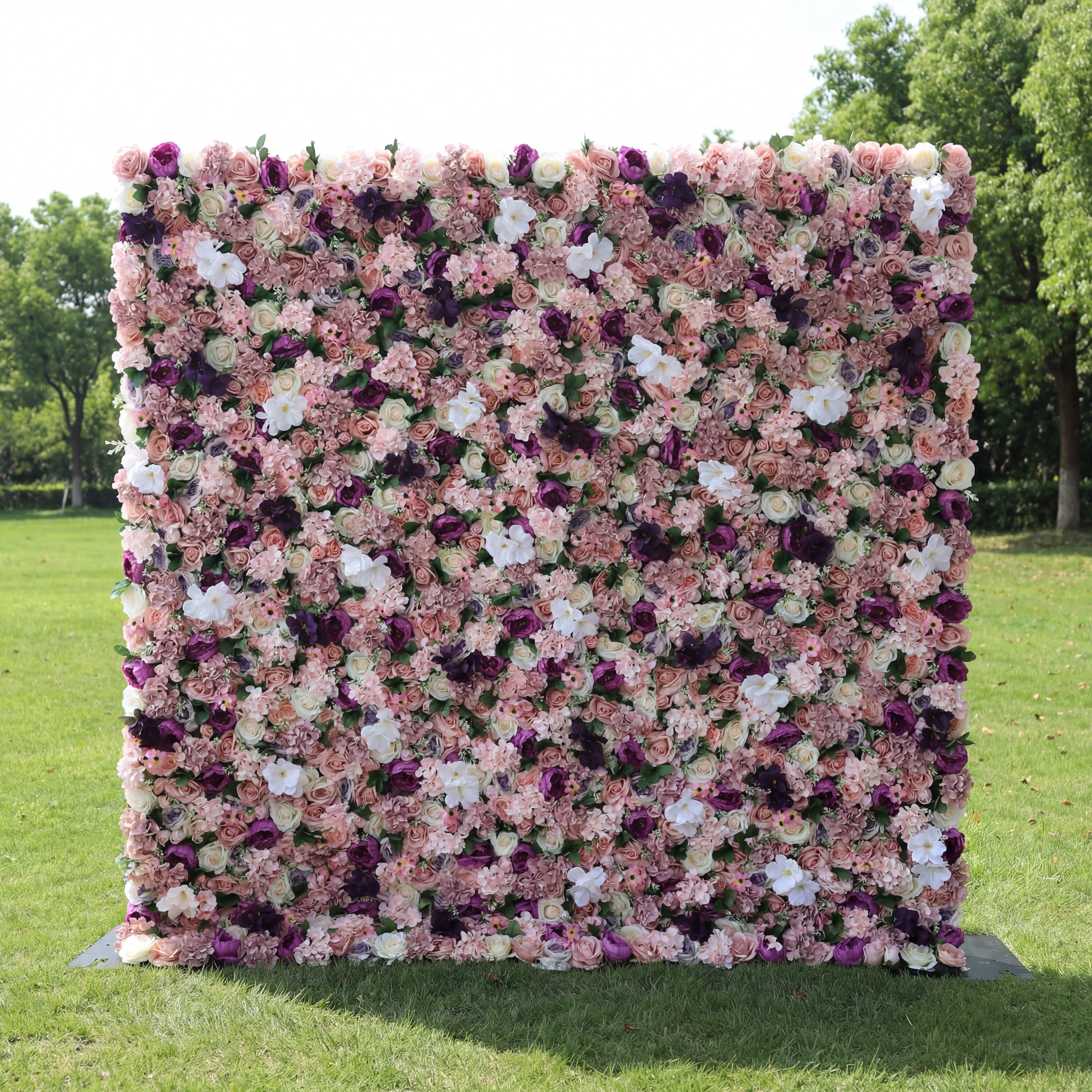 This romantic outdoor floral backdrop features a dense, layered arrangement of artificial blooms in a rich palette of blush, dusty rose, deep plum, and soft white. The lush mix of roses and filler flowers creates a velvety, textured surface that exudes vintage elegance and warmth. Set on a sunlit green lawn framed by trees, it serves as a stunning focal point for weddings, photoshoots, or upscale events.