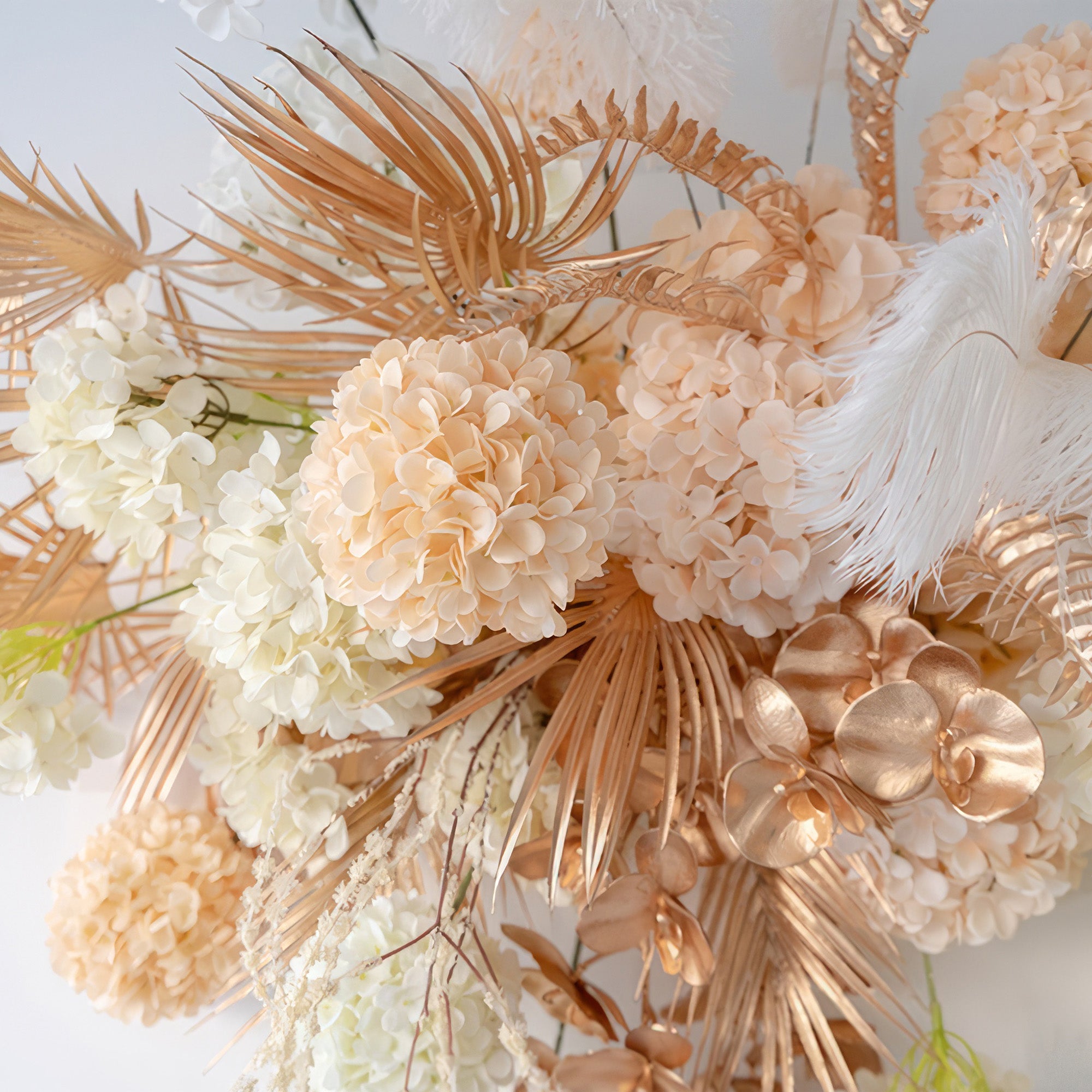 The image displays an artificial floral composition with hydrangeas and orchids, along with palm - like fronds and feathers. In shades of white, peach, and metallic gold, it has a lavish, modern style.