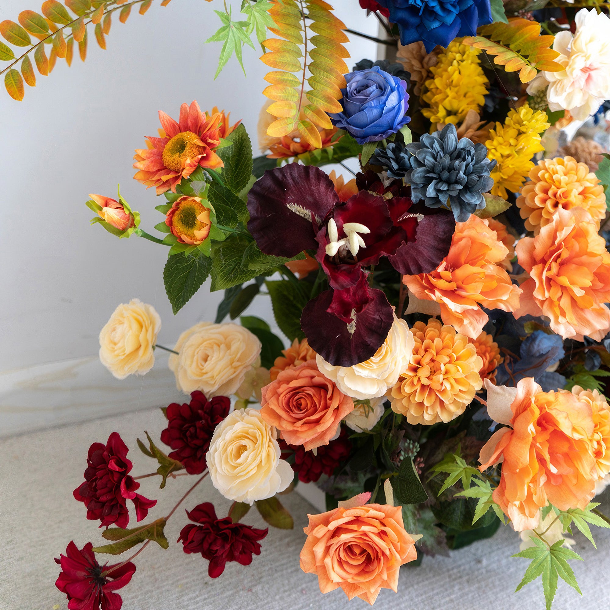 This image displays a colorful artificial flower arrangement. Colors range from deep red and blue to soft cream and bright orange. It includes roses and dahlias. The style is rich and vibrant, exuding a festive atmosphere.