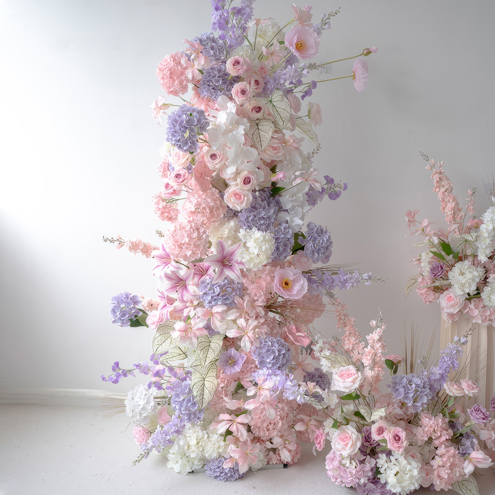 This is a stunning set of floral displays in a soft pastel color scheme. The arrangements feature a mix of pink, lavender, and white artificial flowers, including roses, hydrangeas, and other delicate blooms. The tall vertical arrangement, along with the smaller ones on the ground, creates a romantic and dreamy atmosphere, making them ideal for wedding ceremonies, bridal showers, or other elegant and feminine - themed events.