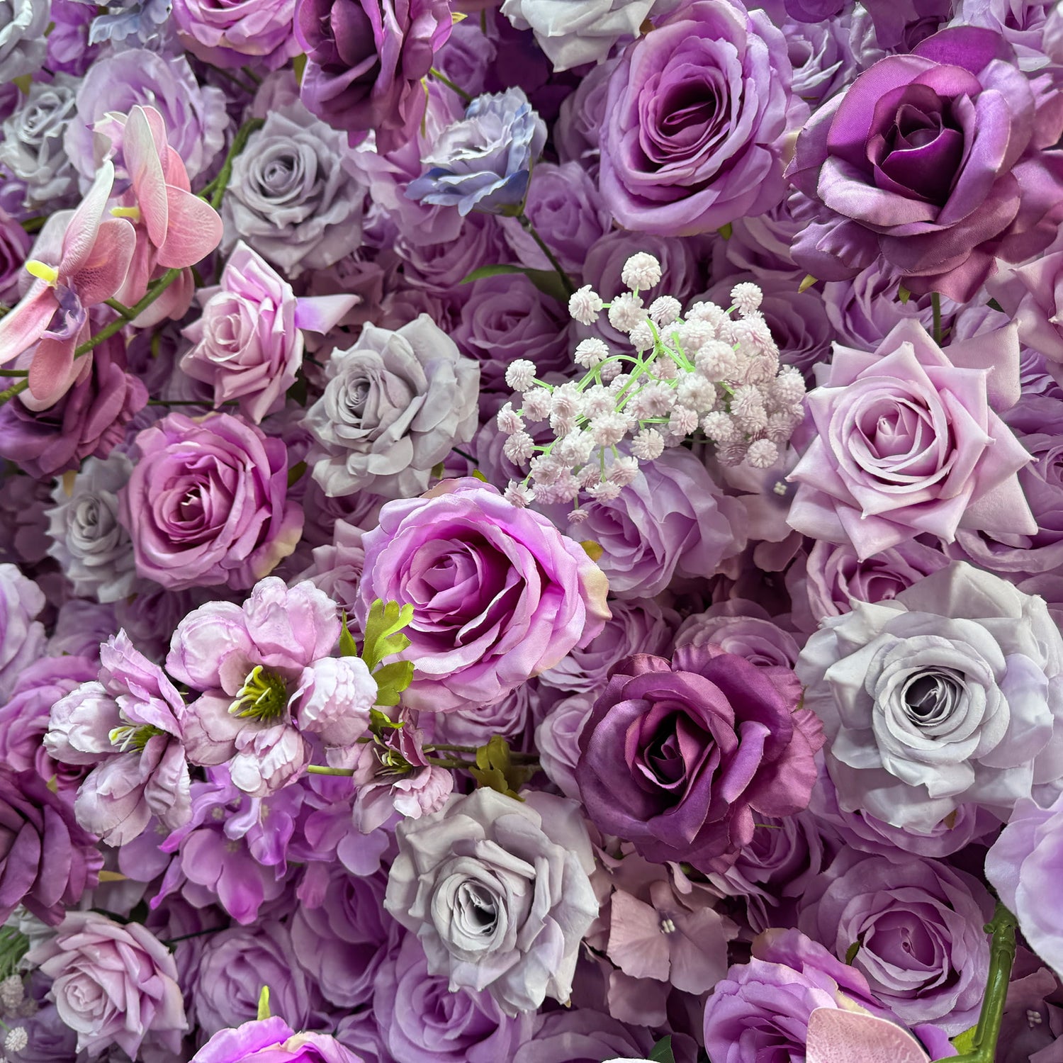 This image showcases a dense arrangement of artificial flowers, predominantly roses in various shades of purple, from light lavender to deep violet. Interspersed are delicate pink orchids and small clusters of white baby&