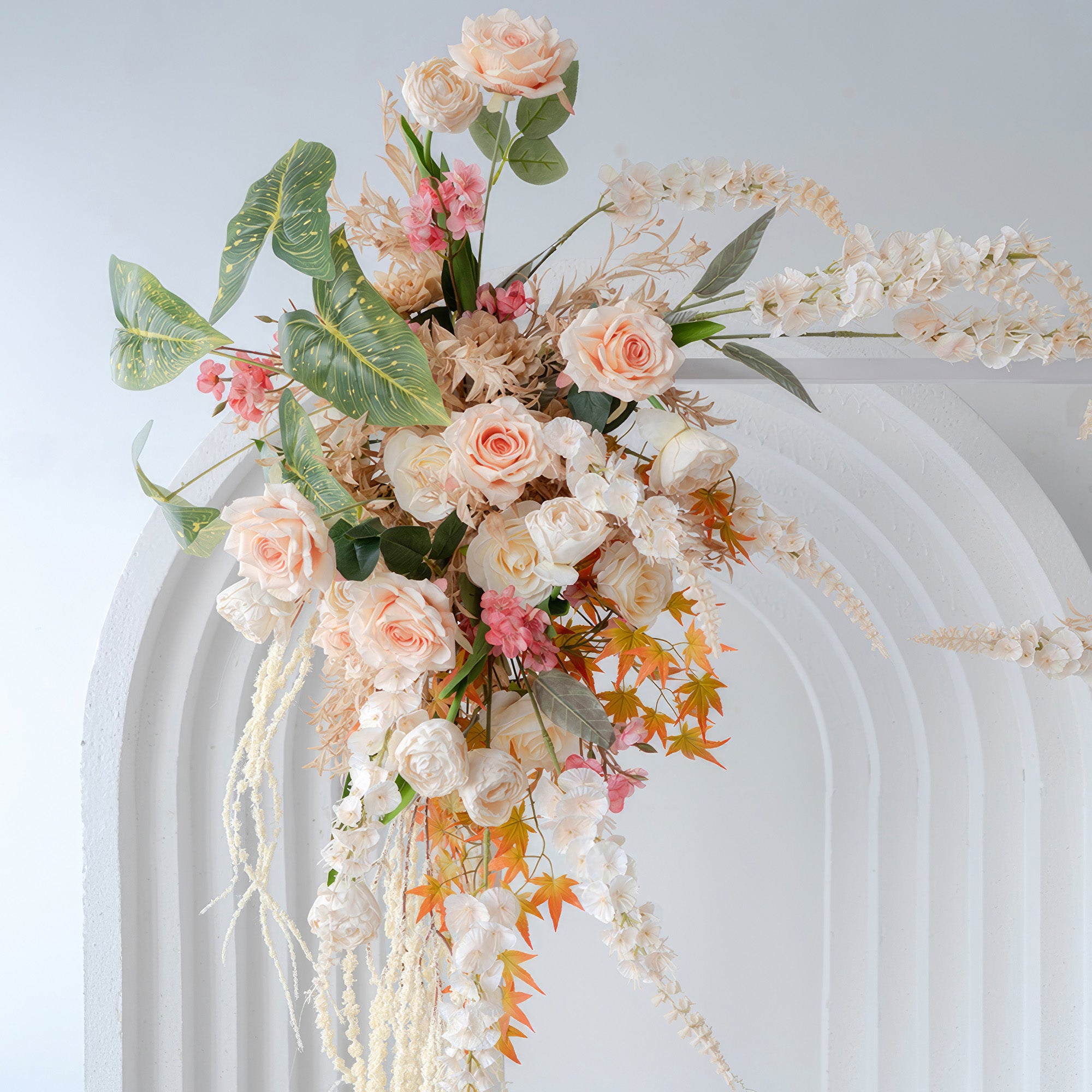 This image features an artificial floral arrangement on an arch, with roses as the main flower. Colors include soft peach, white, pink, and touches of orange. The style is romantic and elegant, suitable for a wedding setting.