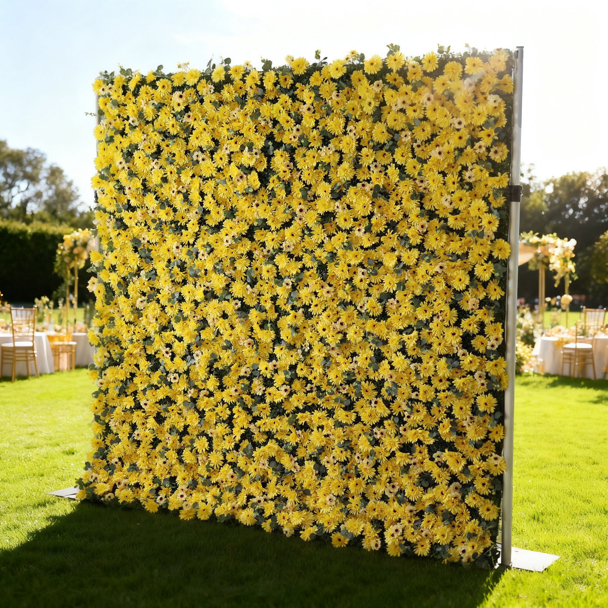 This image showcases a vibrant floral wall by Valar Flowers, set outdoors on a lush green lawn. It features a dense arrangement of artificial yellow flowers, creating a bright and cheerful display. The backdrop is perfect for events, with the sunny hue and lush texture adding a lively touch, complemented by the outdoor setting and event furniture in the background.
