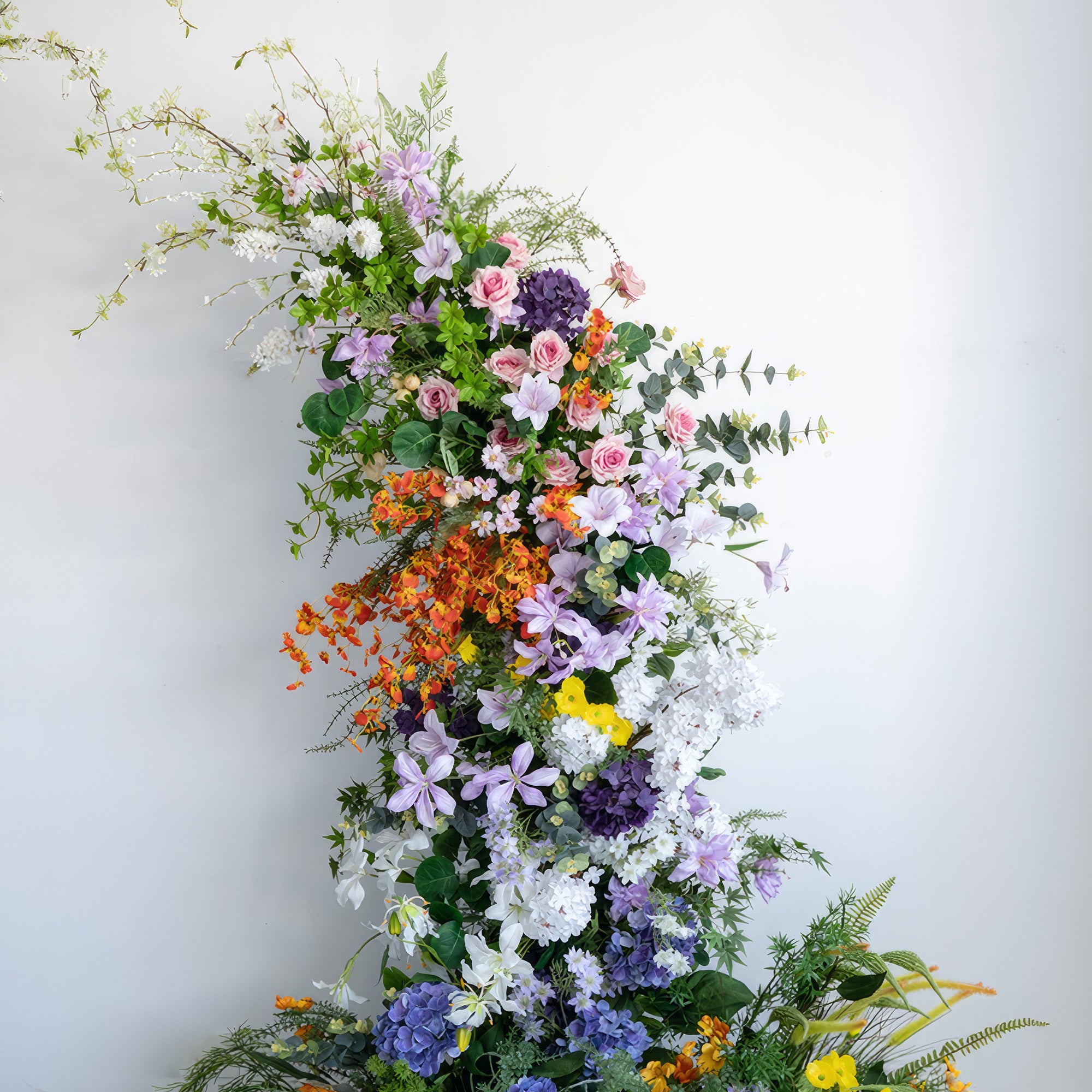 This is an artificial floral installation with a vivid color scheme. It combines orange, purple, pink, white, and yellow. Flowers like hydrangeas, roses, and other assorted blooms are used, creating a lush, dynamic, and nature - evoking style.