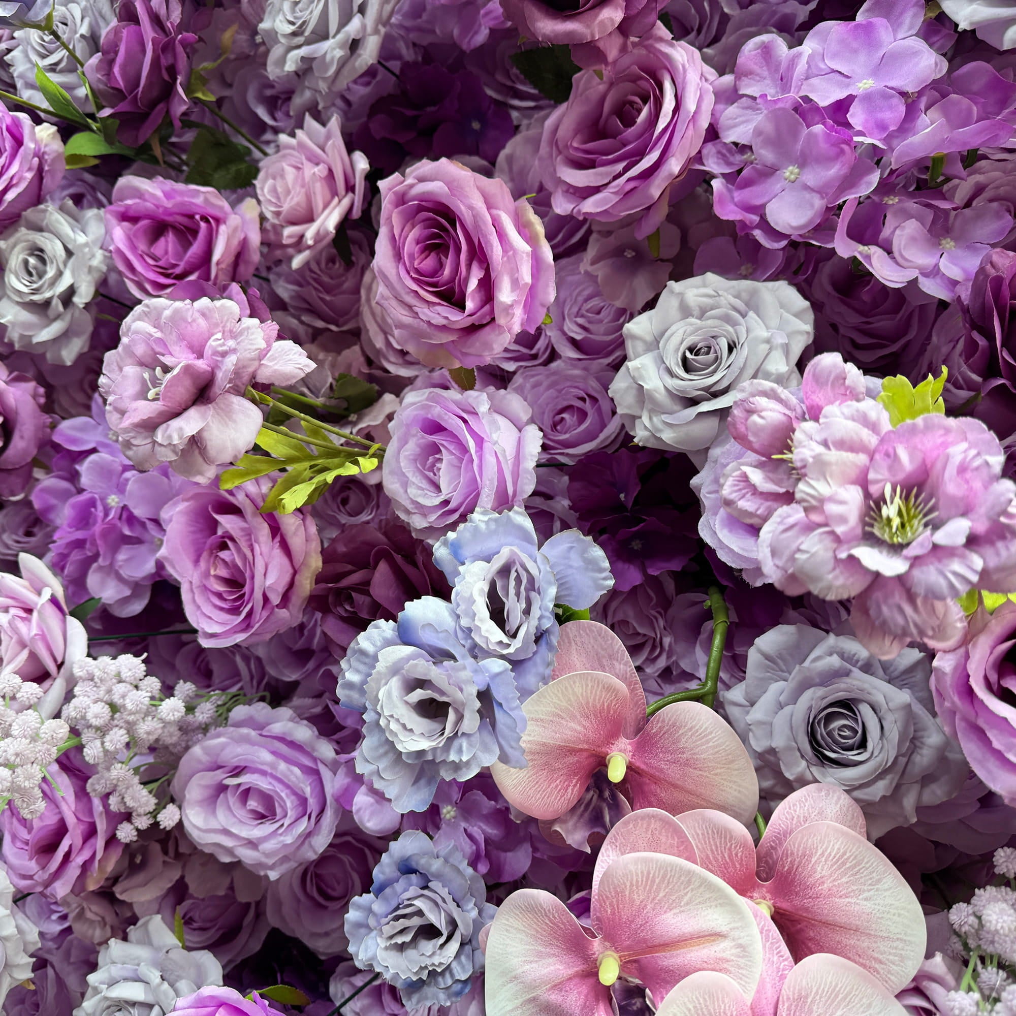 This image showcases a lush arrangement of artificial flowers. Prominent among them are roses in various shades of purple, lavender, and pink, along with delicate orchids and clusters of small white flowers. The color palette is a harmonious blend of cool purples and soft pinks, creating a romantic and elegant style, ideal for event decorations.
