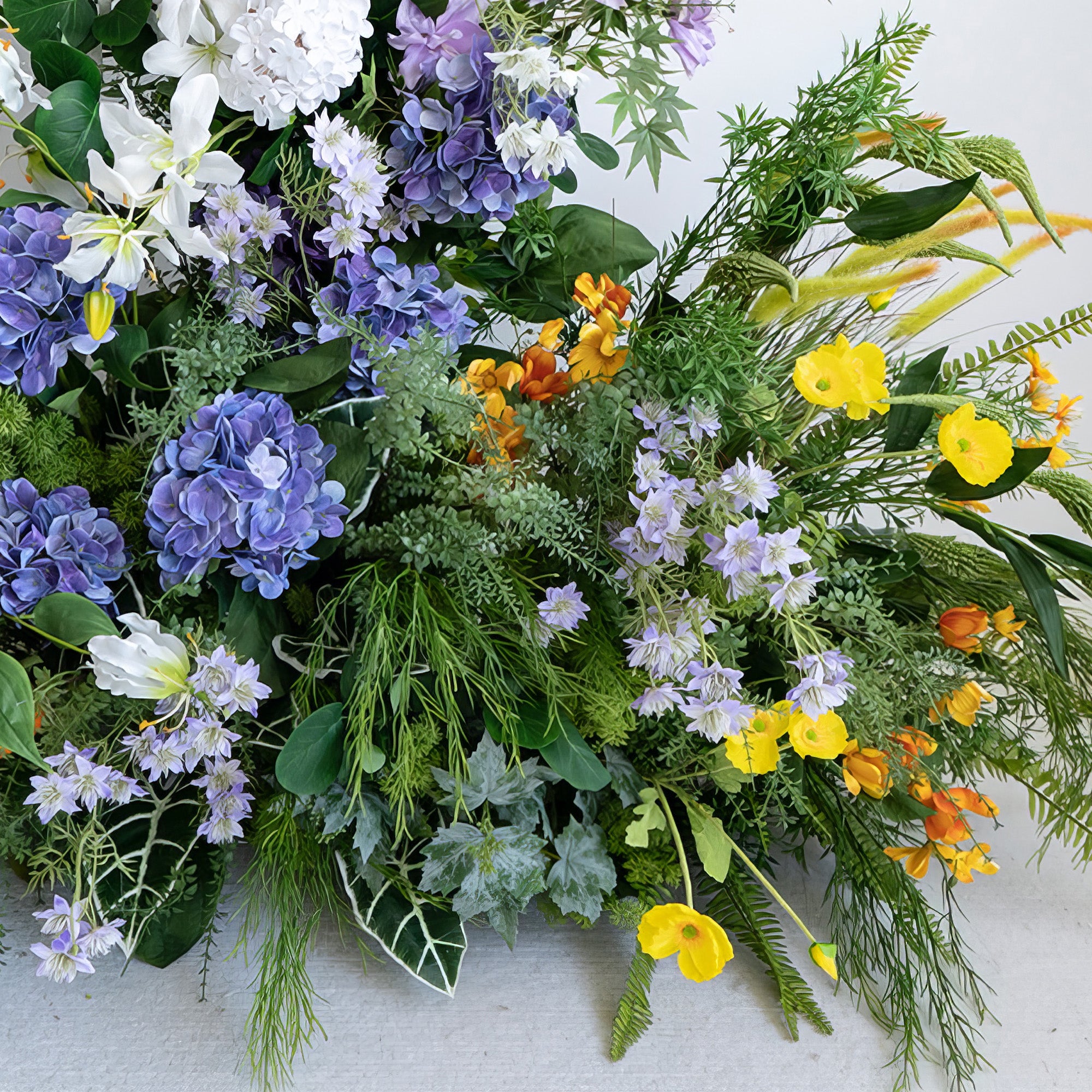 This image displays a vibrant artificial flower arrangement. The colors include deep purple, white, yellow, and orange, with a backdrop of various greens. It features hydrangeas, along with smaller purple and yellow blooms. The style is lively and nature - inspired, creating a rich and textured look.