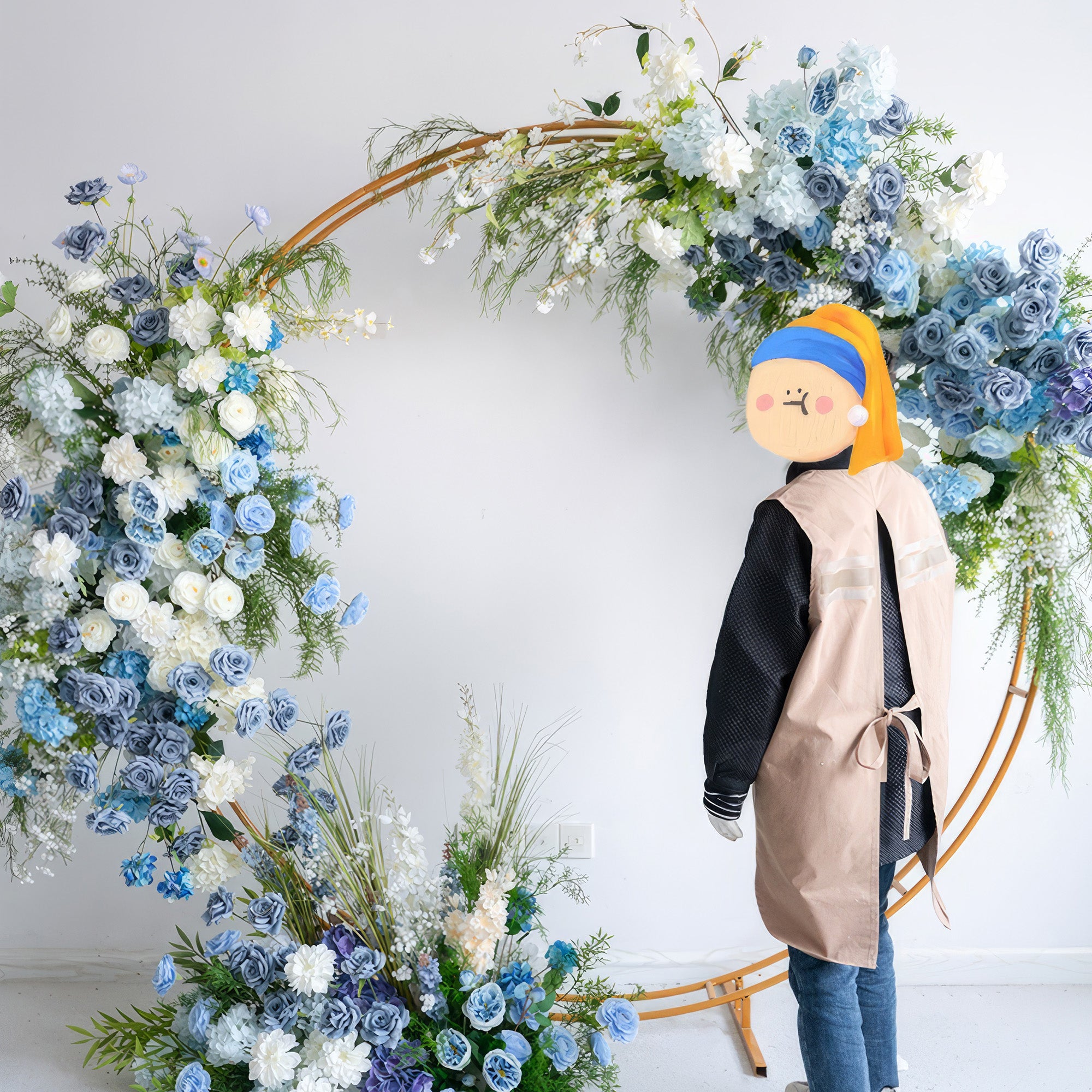 A sleek, gold circular floral arch from Valar Flowers, adorned with a serene palette of sky blue, navy, and creamy white blooms—including hydrangeas, roses, and delicate orchids—intertwined with cascading greenery. A person in a beige apron stands beside it, creating a sophisticated, romantic atmosphere perfect for a luxury wedding or special celebration.