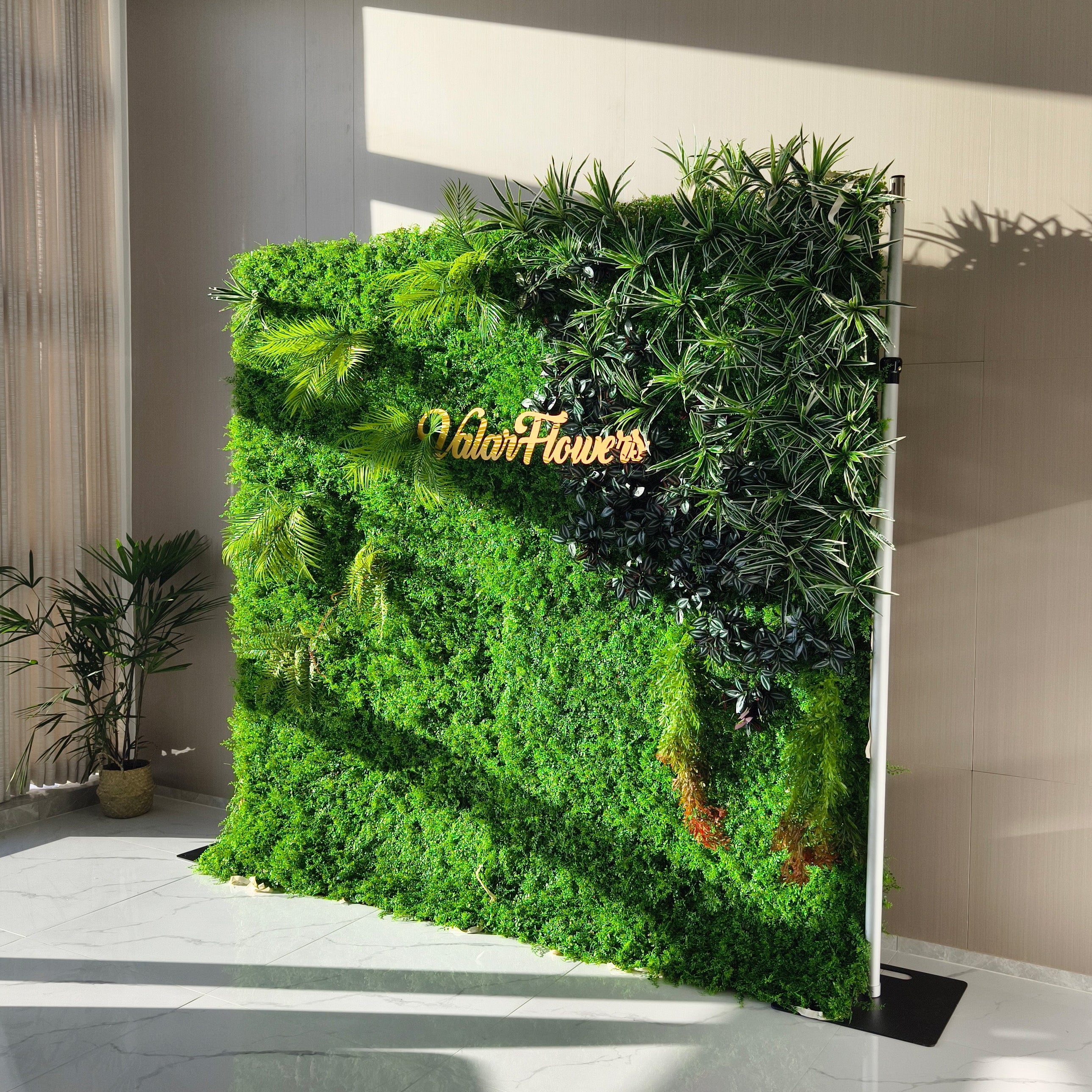 This image showcases a lush artificial greenery wall by ValarFlowers, set indoors on a glossy marble floor. It features a dense base of vibrant green foliage, artfully accented with varied plants like palm fronds, textured greens, and variegated leaves. The prominent “ValarFlowers” logo adds a branded touch. Natural light casts shadows, enhancing the realistic textures, while a potted plant and modern interior elements complement the scene, exuding a serene and sophisticated atmosphere ideal for events.