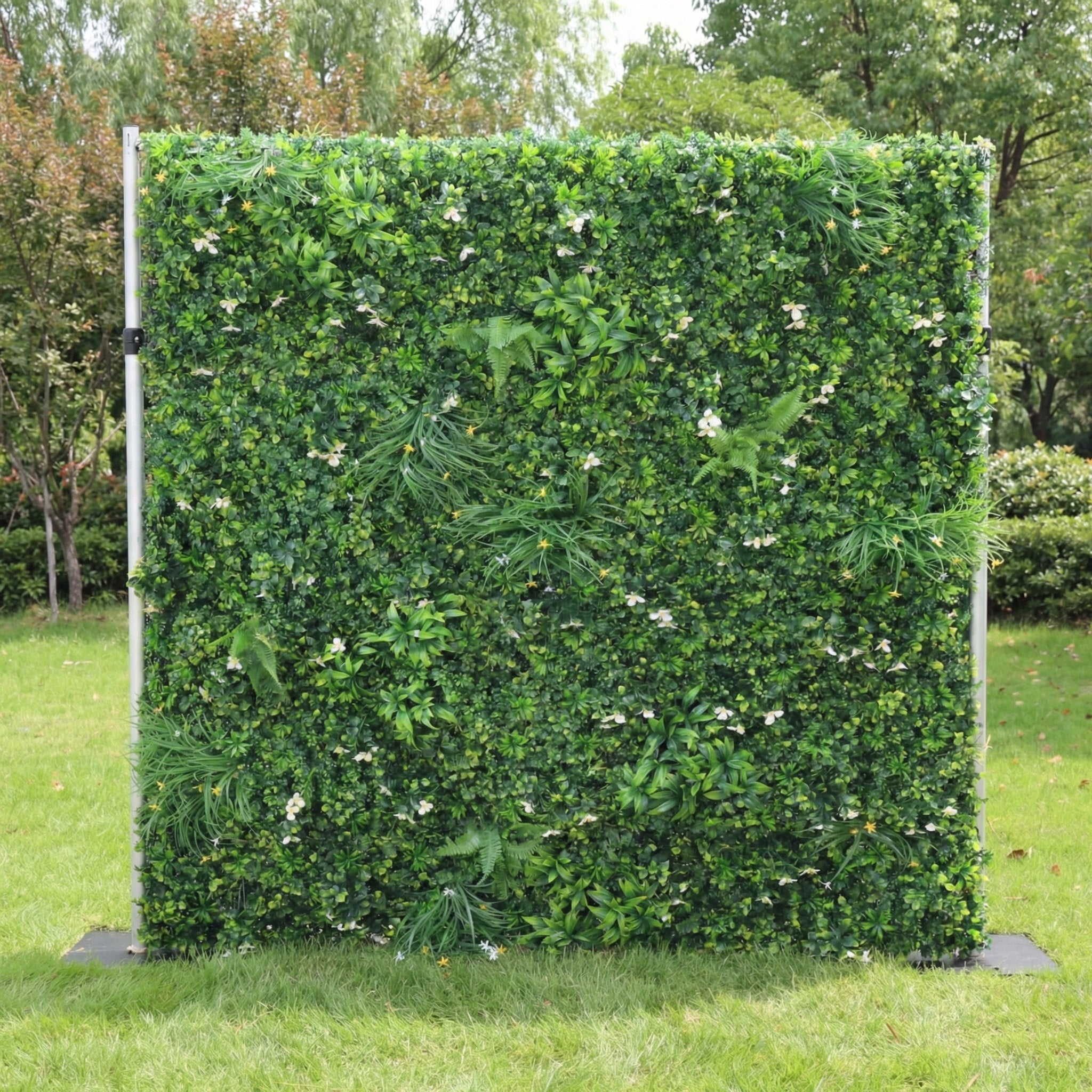 This outdoor greenery wall from Valar Flowers is a lush, textured backdrop of dense, vibrant green foliage—including ferns, leafy shrubs, and grassy accents—interspersed with delicate white and yellow wildflowers. Set on a sunlit green lawn framed by leafy trees, the design evokes a fresh, natural, and elegantly understated atmosphere ideal for weddings, photo backdrops, or elegant outdoor celebrations.