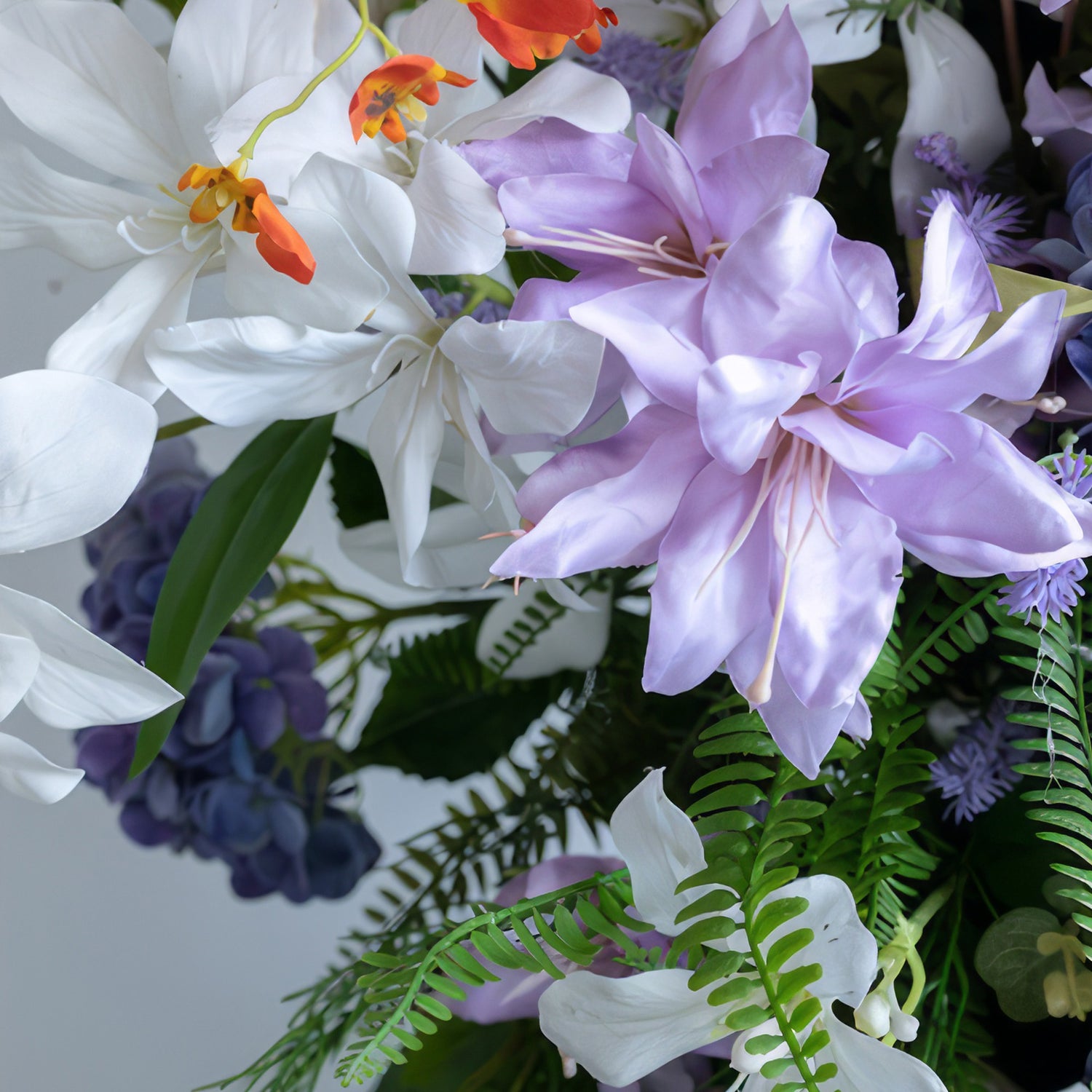 This close - up shows an artificial flower arrangement. Colors are white, purple, and orange, with green foliage as a backdrop. The flowers include large, broad - petaled white and purple blooms, along with small orange ones, presenting a fresh and colorful style.