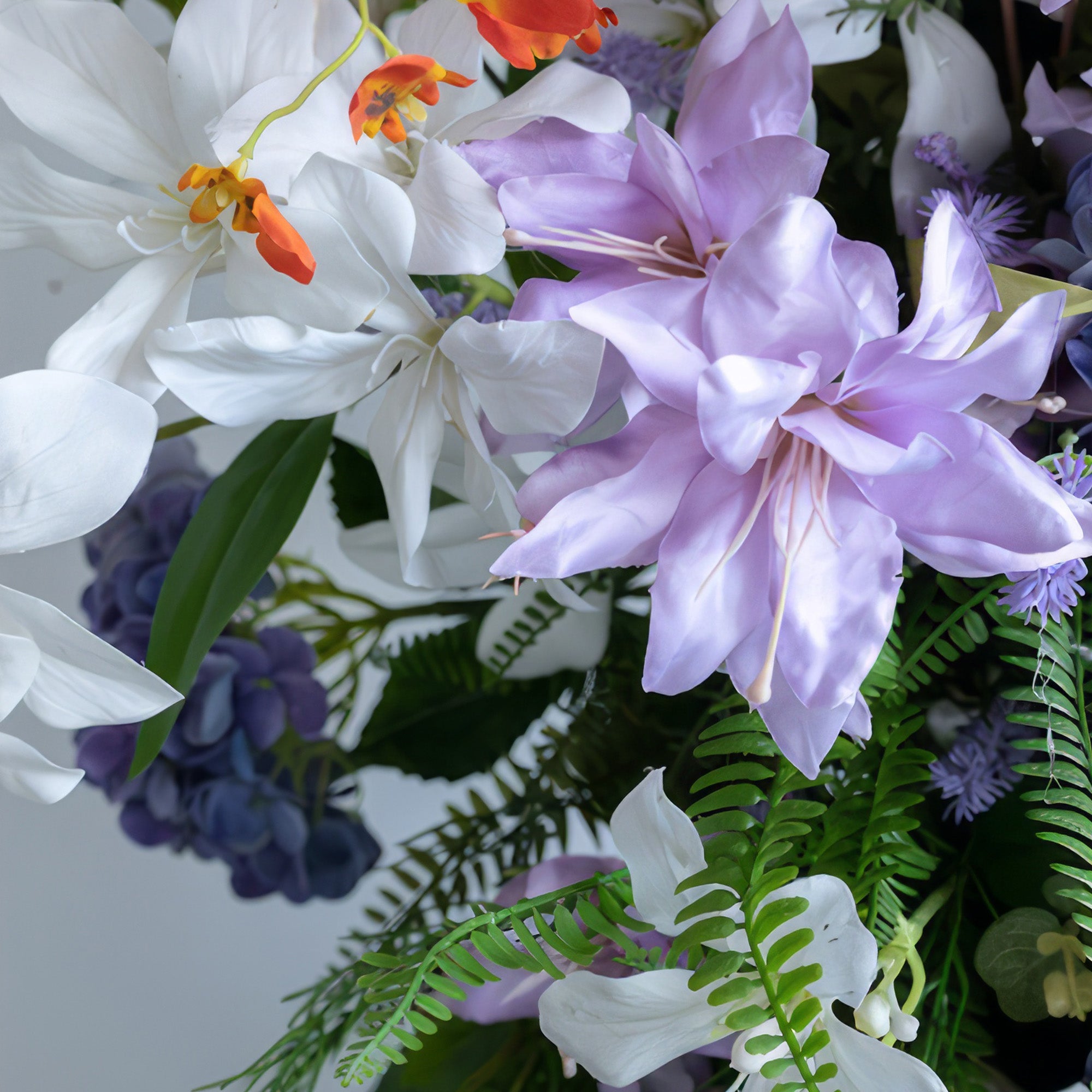 This close - up shows an artificial flower arrangement. Colors are white, purple, and orange, with green foliage as a backdrop. The flowers include large, broad - petaled white and purple blooms, along with small orange ones, presenting a fresh and colorful style.