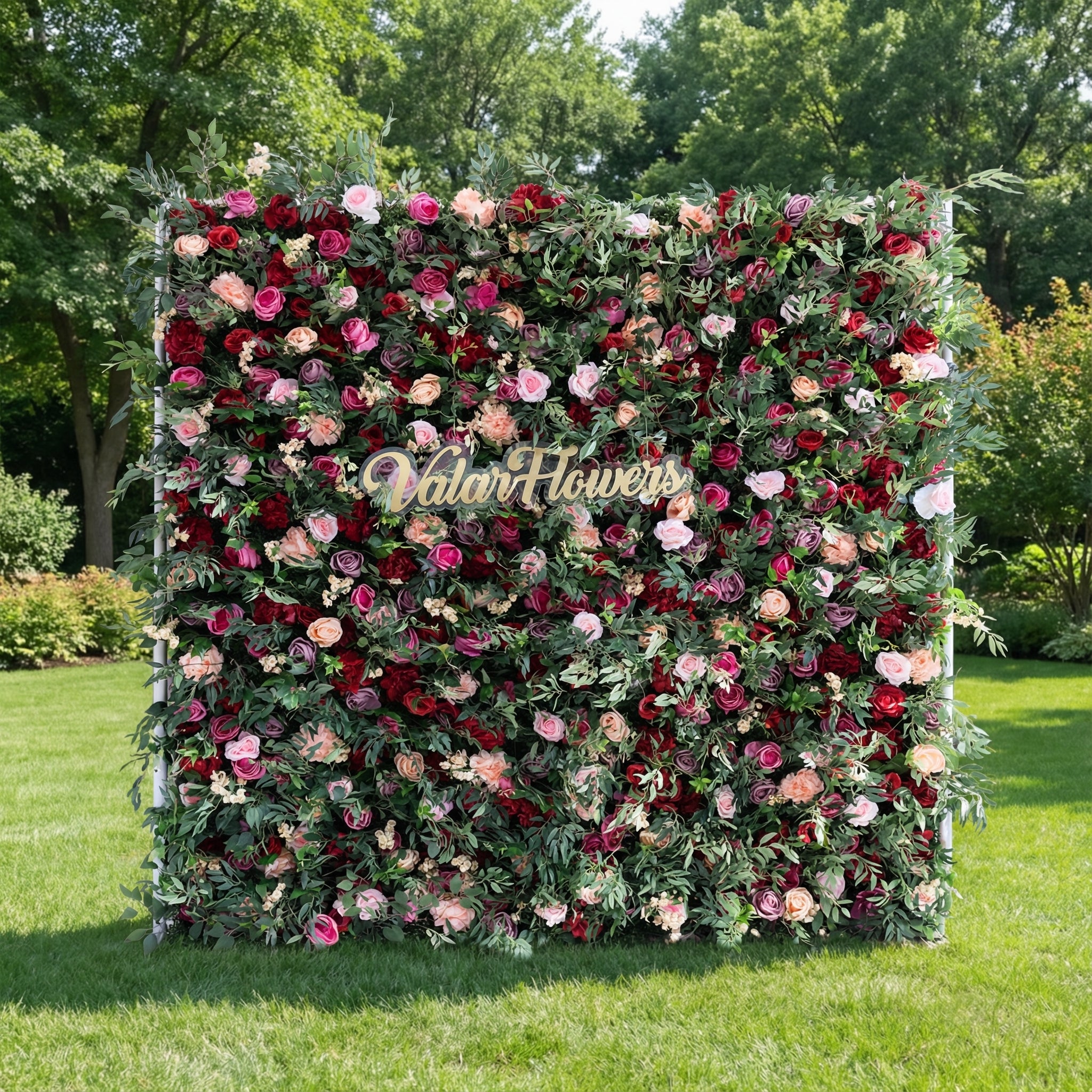 This image showcases a vibrant flower wall by Valar Flowers, featuring a dense arrangement of artificial roses in rich reds, pinks, and peaches, complemented by green foliage. Set outdoors on lush grass with trees in the background, a gold “Valar Flowers” sign is integrated at the center. It exudes elegance and sophistication, ideal for events, highlighting the brand’s craftsmanship in creating eye-catching floral decor.