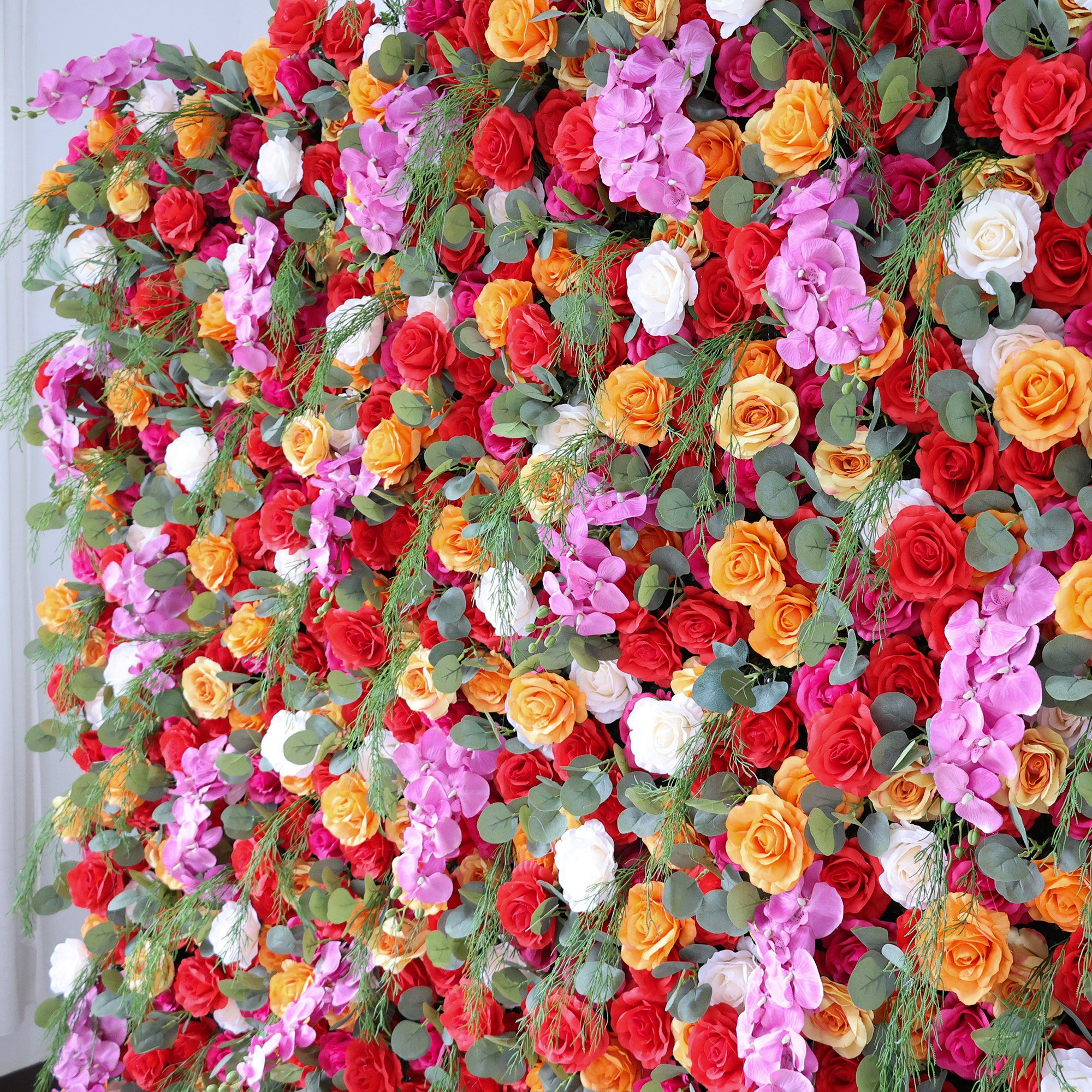 This image showcases a vibrant floral arrangement by Valar Flowers. It features a dense cluster of artificial flowers in a rich palette of red, orange, pink, and white, interspersed with lush green foliage. The blooms, including roses and other complementary flowers, are intricately arranged to create a lush, textured, and visually striking display. Perfect for adding a touch of energy and elegance to events or spaces, the arrangement exudes a lively and sophisticated atmosphere.