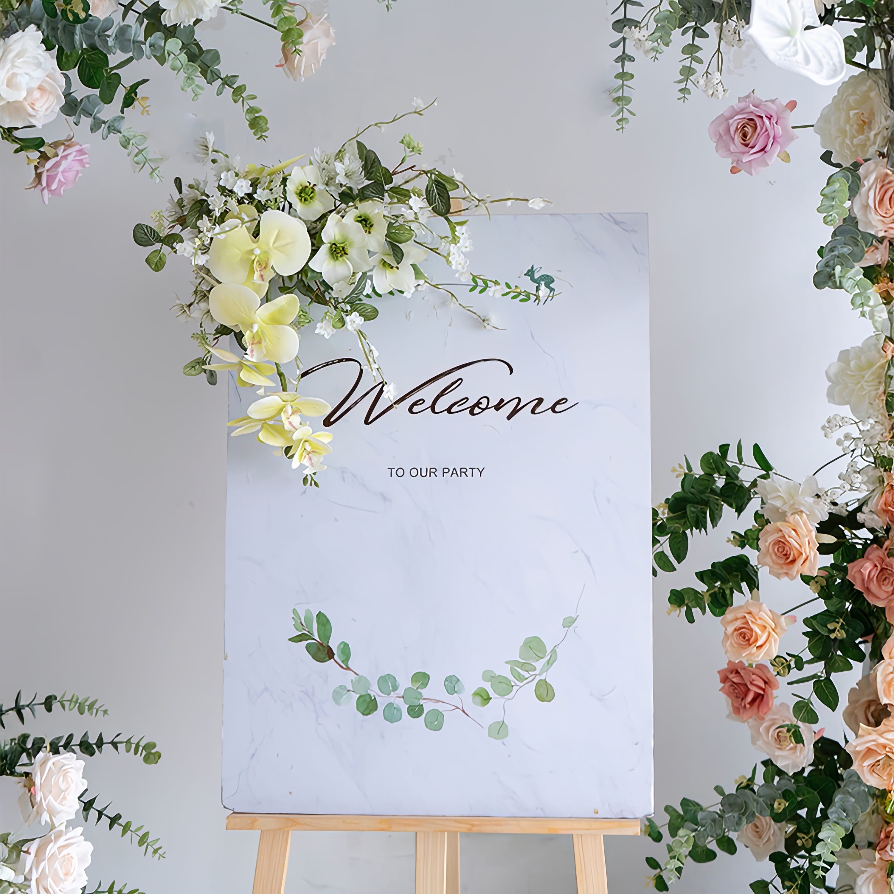 A "Welcome to Our Party" sign on a wooden easel, framed by a lush floral arrangement from Valar Flowers. The sign features a marble-patterned background with delicate eucalyptus illustrations, while the surrounding blooms include blush pink roses, creamy white orchids, and silvery-green eucalyptus, creating an elegant, inviting aesthetic ideal for weddings, parties, and special events.