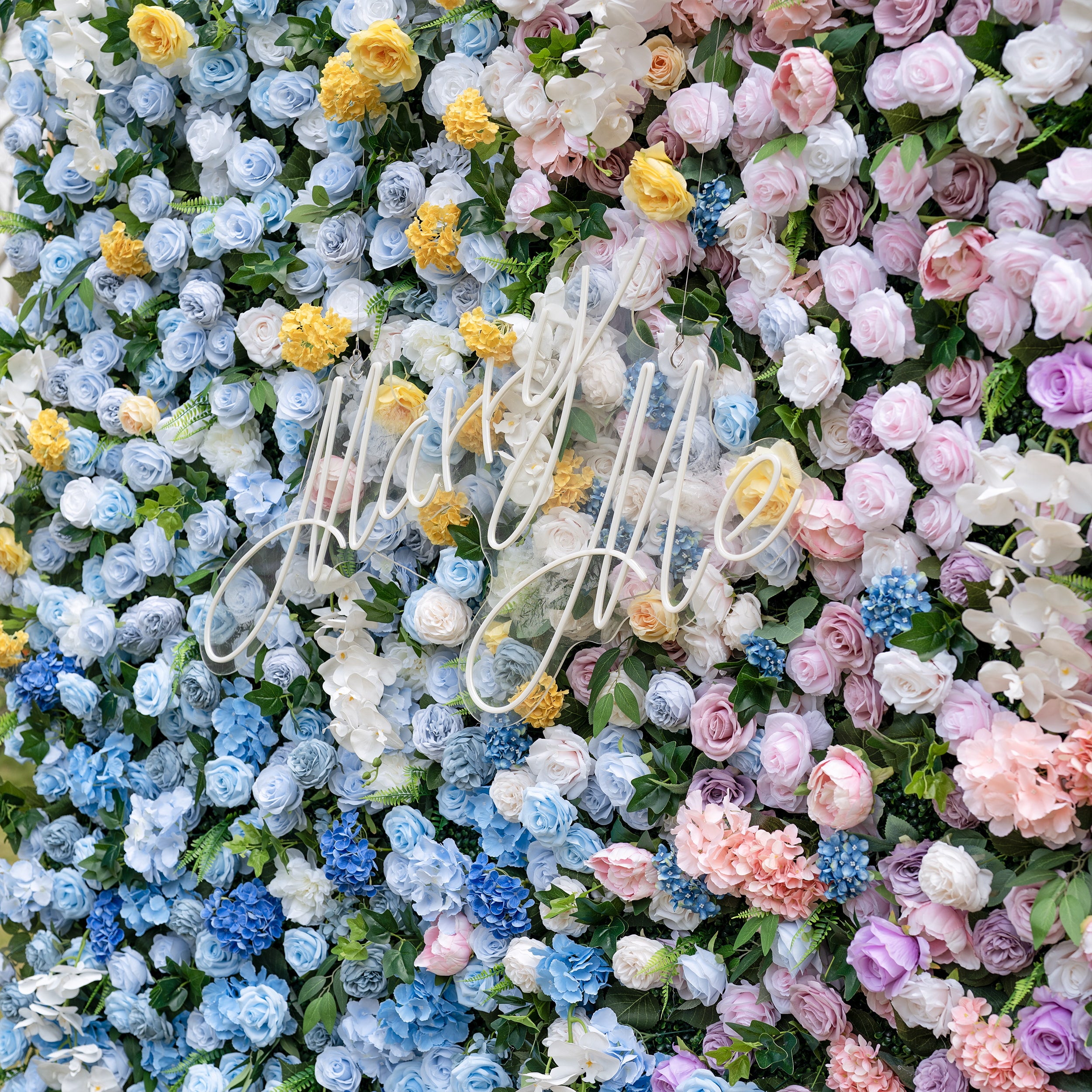 This is a stunning gradient artificial flower wall backdrop, centered with an elegant "Marry Me" neon script sign. It features a lush, dense arrangement of lifelike blooms: soft sky-blue delphiniums at the bottom, transitioning upward to creamy white roses, sunny yellow poppies, blush pink peonies, and lavender roses. This premium, reusable floral display creates a romantic, showstopping focal point ideal for wedding proposals, ceremonies, photo booths, and luxury event decor.