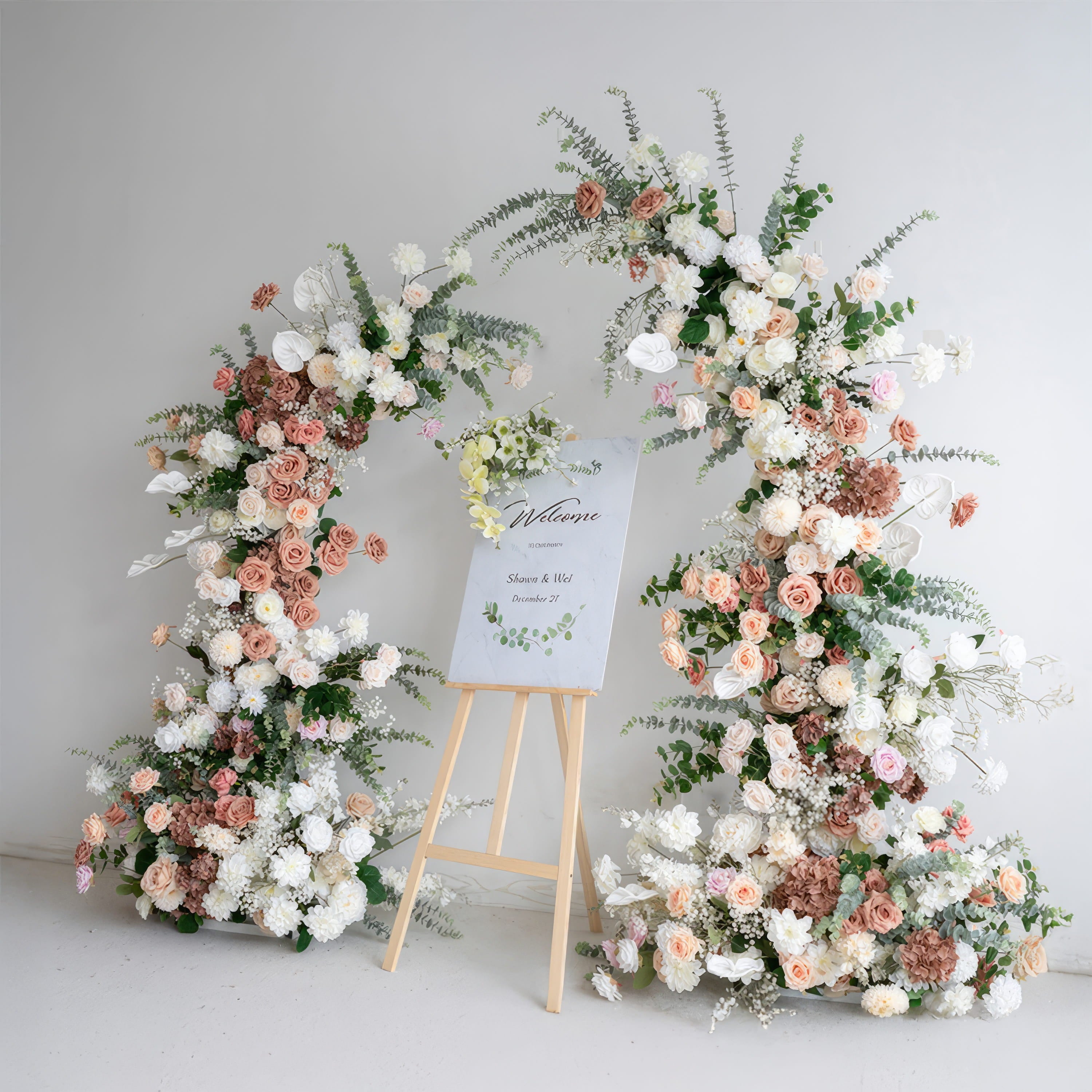 A khaki color floral archway with artificial flowers, set against a white background, with a wooden sign in front displaying &