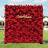 This image showcases a large, square floral wall densely covered with vibrant red roses. "Valar Flowers" is displayed in golden text. Set outdoors on a lush green lawn under a bright blue sky with scattered clouds, and with white tents and floral arrangements in the background, it creates a striking and elegant atmosphere, perfect for events or decorative displays.