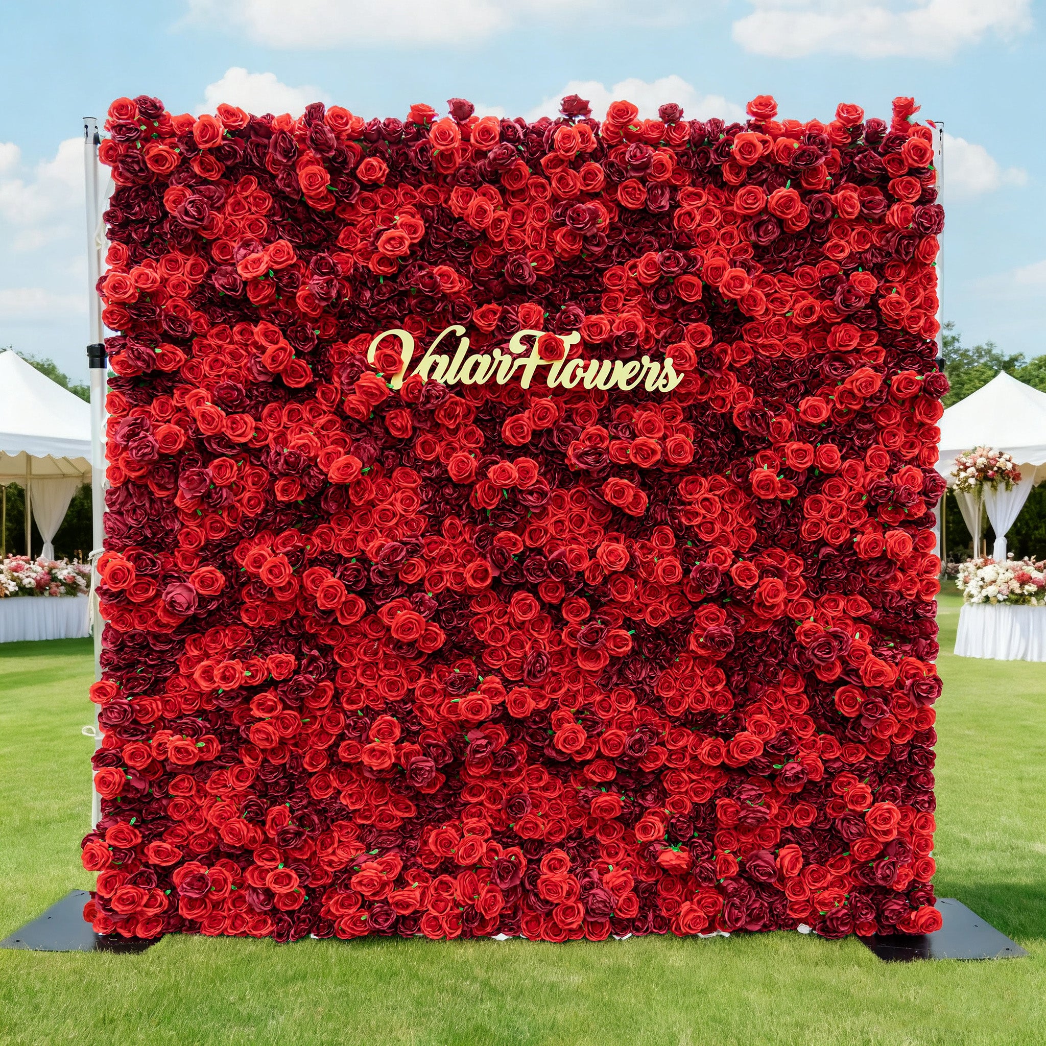 This image showcases a large, square floral wall densely covered with vibrant red roses. "Valar Flowers" is displayed in golden text. Set outdoors on a lush green lawn under a bright blue sky with scattered clouds, and with white tents and floral arrangements in the background, it creates a striking and elegant atmosphere, perfect for events or decorative displays.